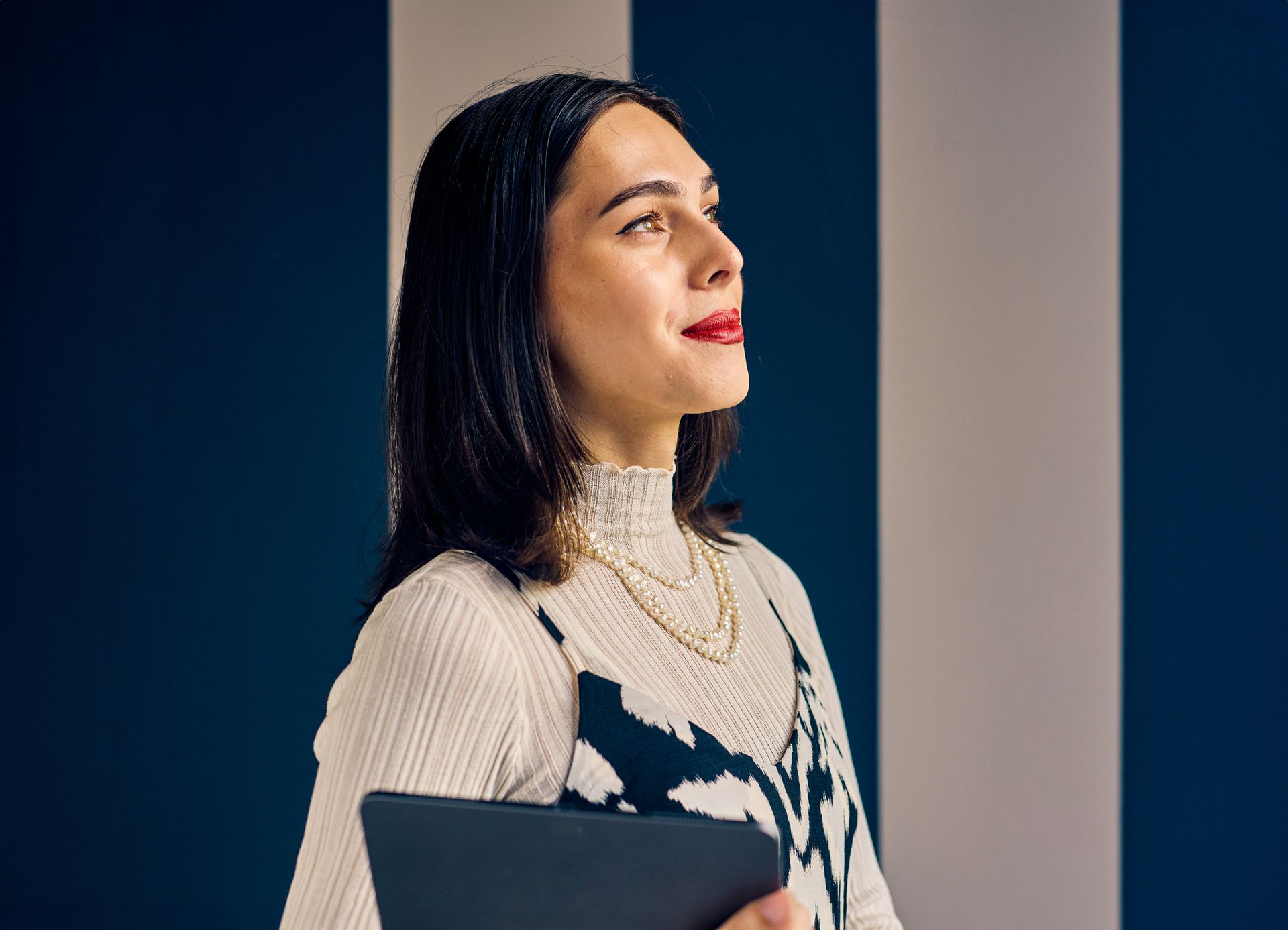 Woman with red lipstick looking up, holding a tablet, against a blue and white striped wall.
