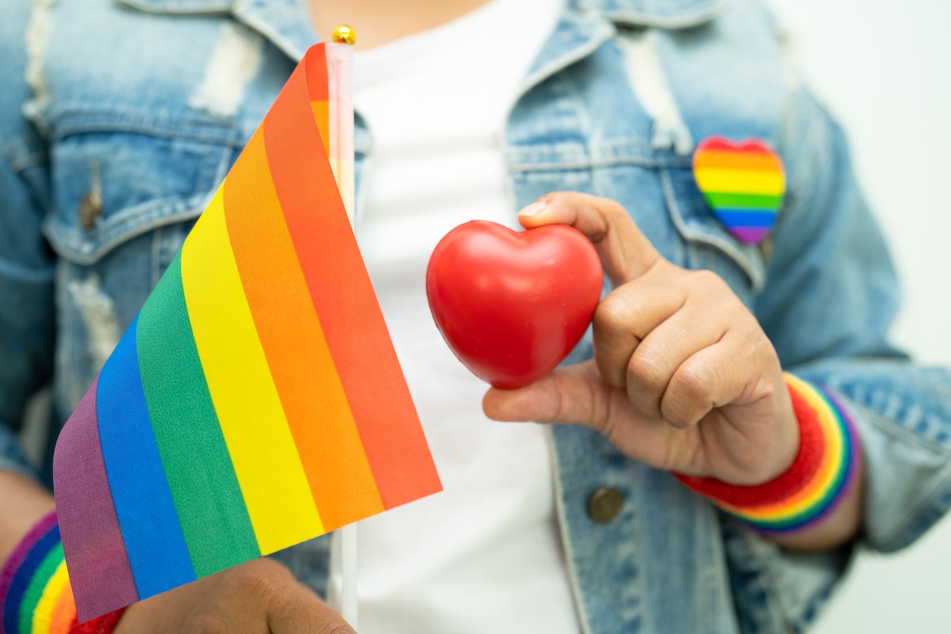 Person holding a rainbow flag and red heart, wearing rainbow wristbands and a denim jacket.