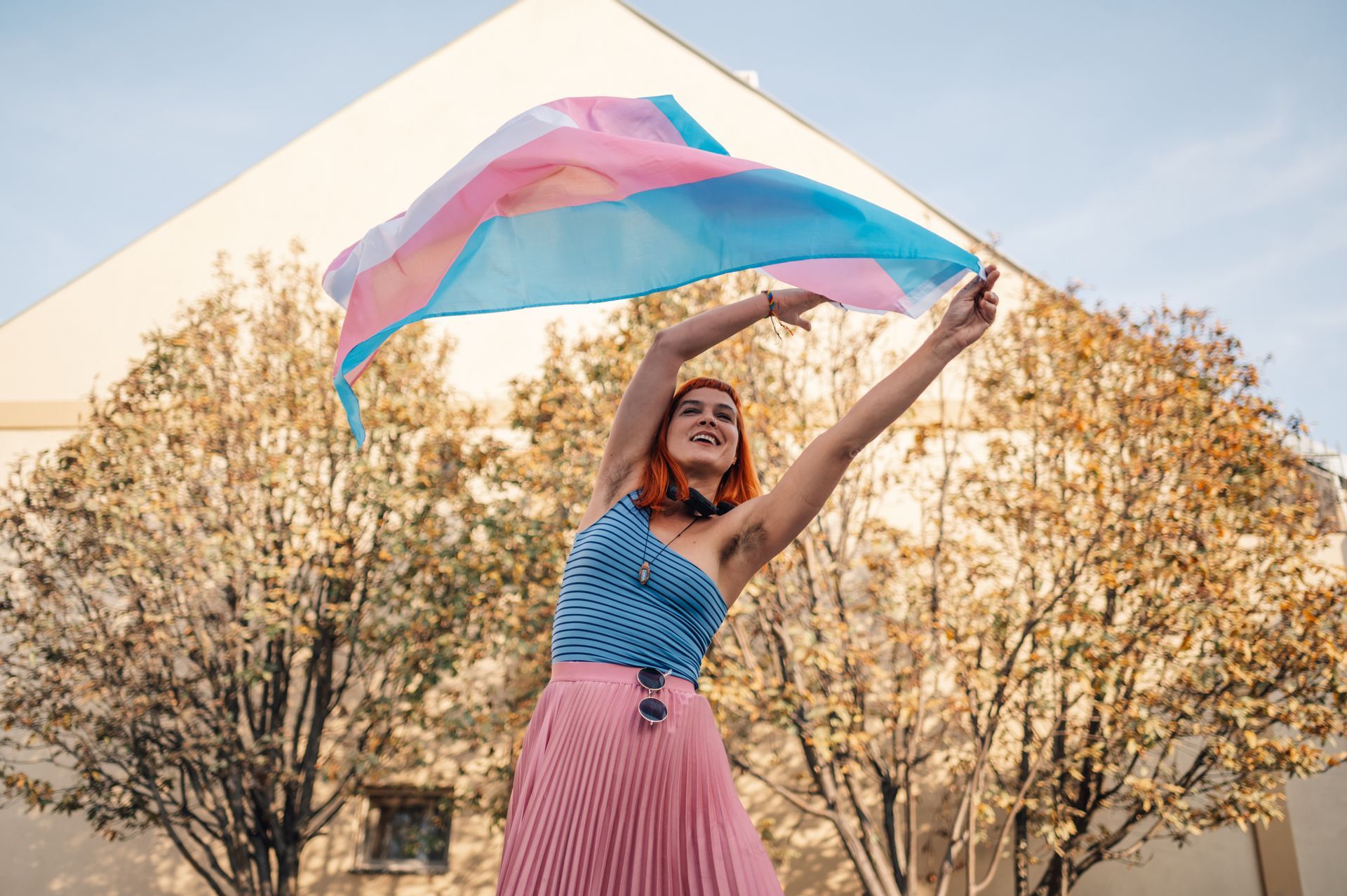 Person with red hair holding a transgender flag. Outdoors, standing in front of building and trees, celebrating.