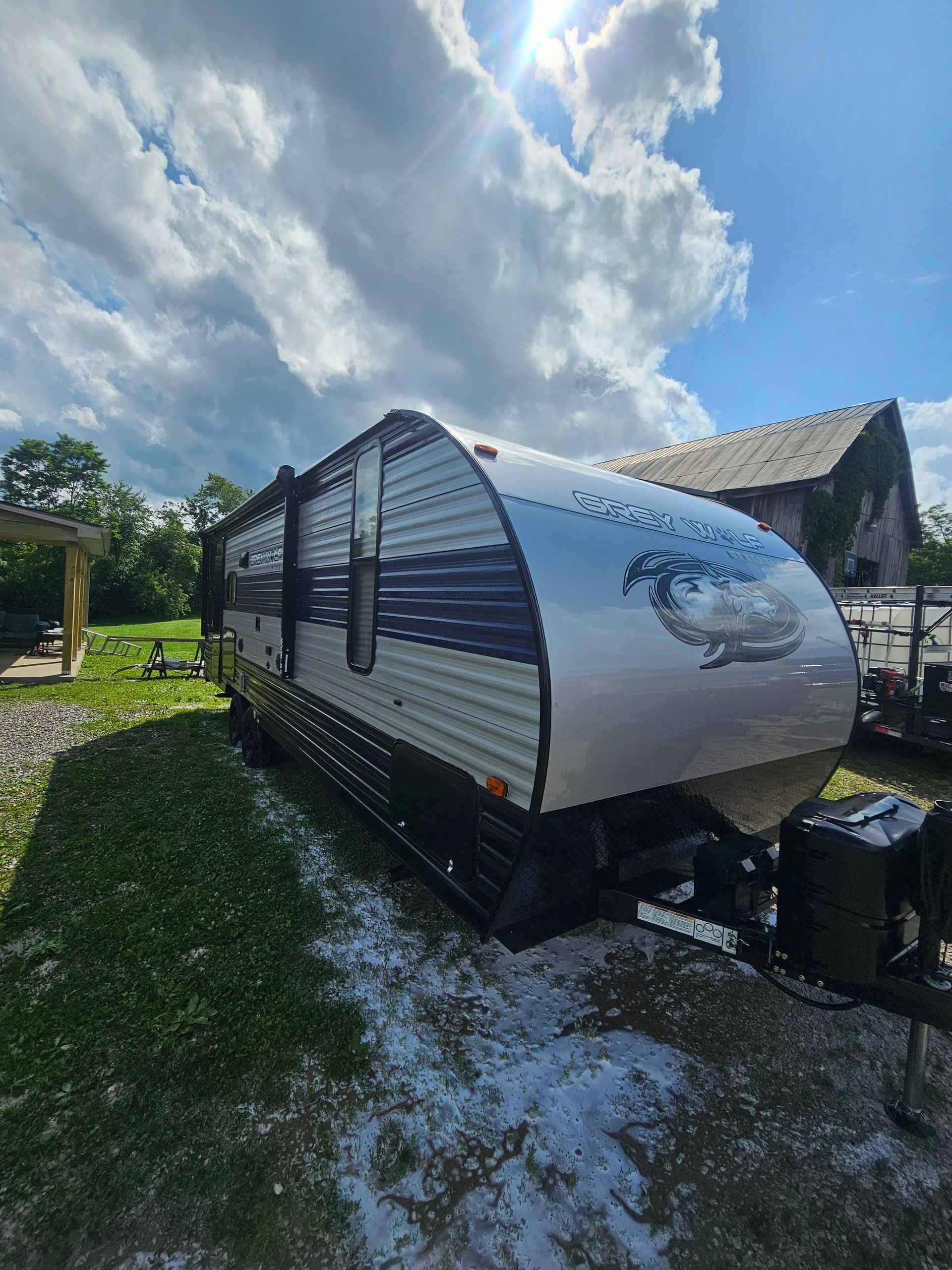 Silver and blue travel trailer parked on grass, with a barn in the background under a cloudy sky.