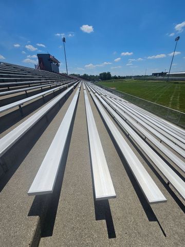 Empty stadium seating on a sunny day. Bleachers face a grassy field, under a blue sky.