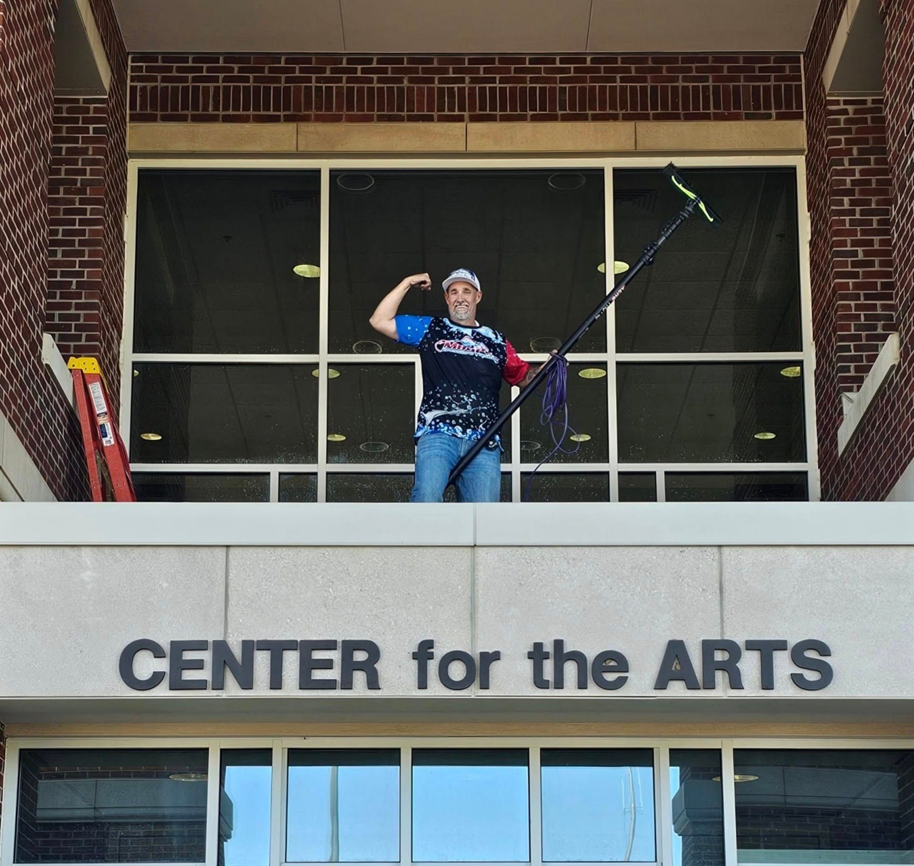 Man power washes a sidewalk in front of a house. Black van parked on street. Trees and grassy yard.