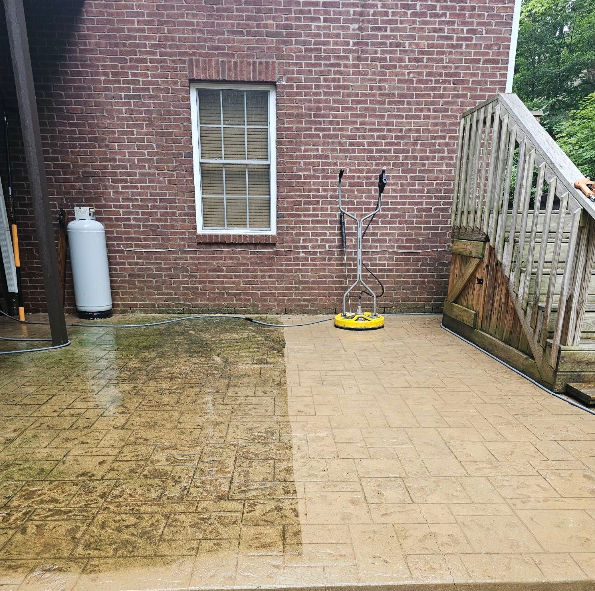 A patio partially cleaned with a pressure washer. Brick building, wooden stairs, and a propane tank in the background.