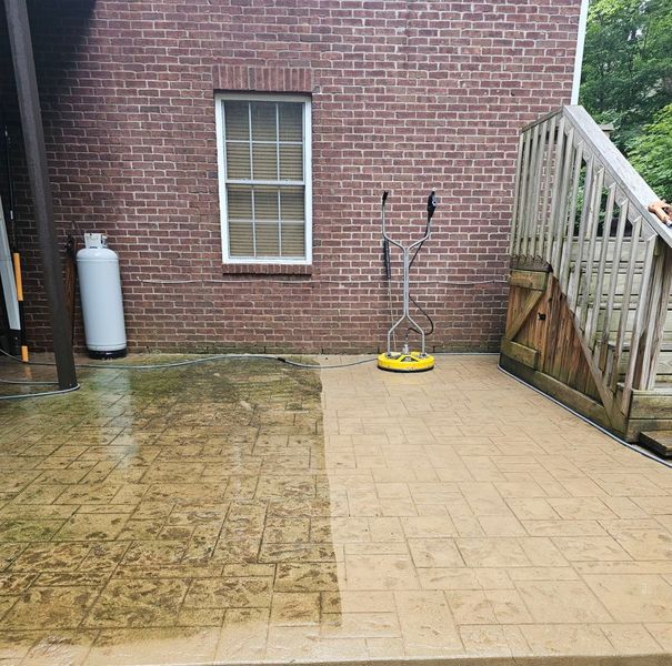 A patio partially cleaned with a pressure washer. Brick building, wooden stairs, and a propane tank in the background.