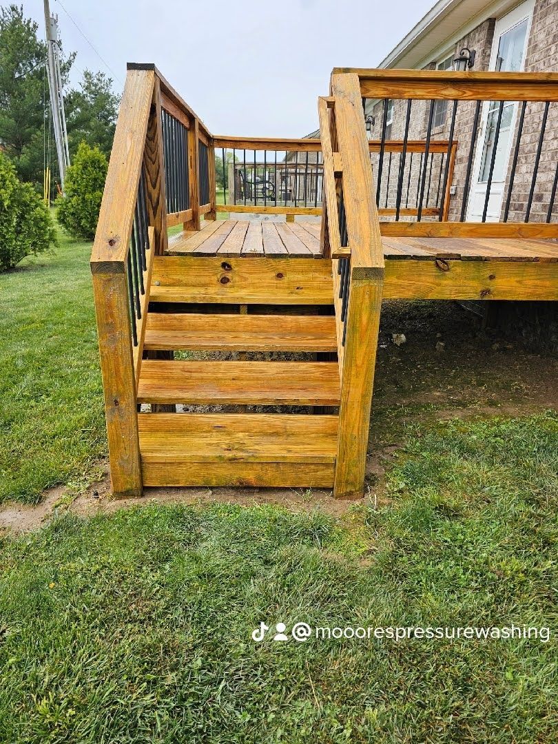 Man power washes a sidewalk in front of a house. Black van parked on street. Trees and grassy yard.