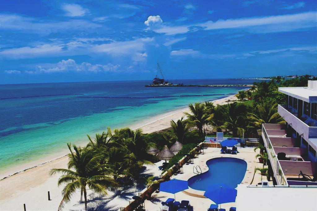 Resort frente al mar con piscina y vista al mar; arena blanca, agua azul, palmeras.