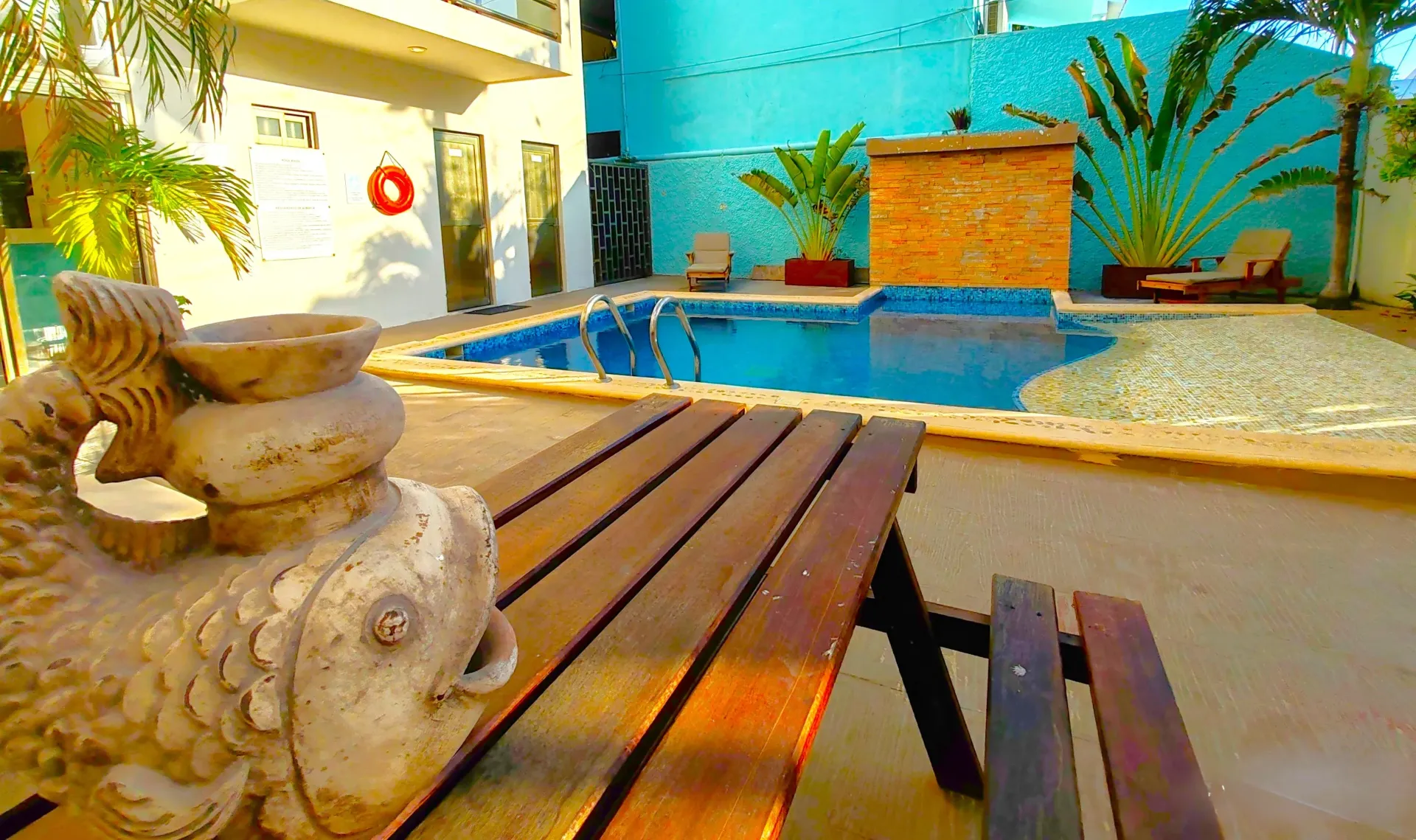 Mesa de madera frente a una estatua de pez, piscina al fondo con pared azul, palmeras y sol.