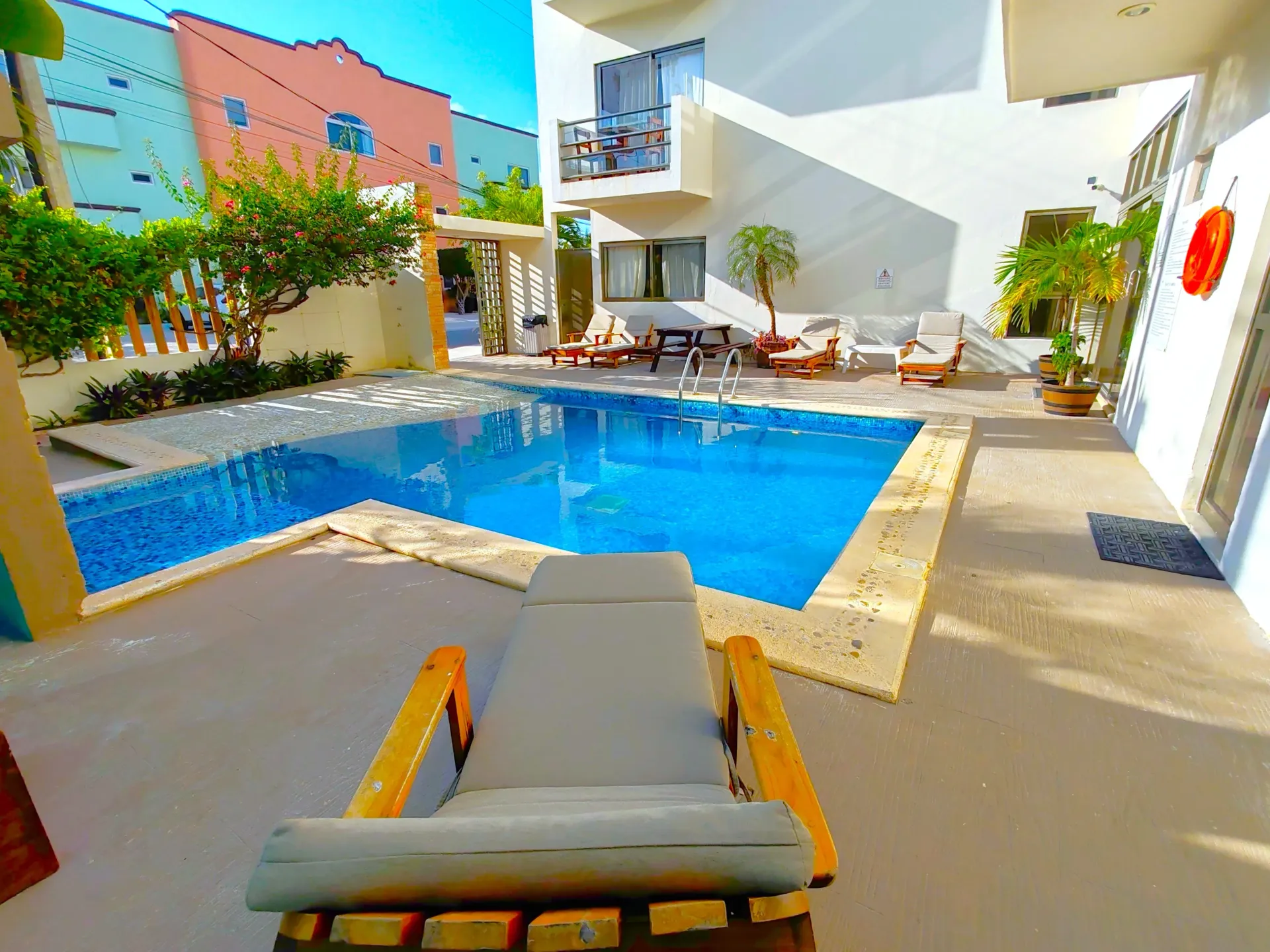 Un sillón frente a la piscina. Edificios blancos rodean la piscina, algunos con balcones. Cielo azul, soleado.