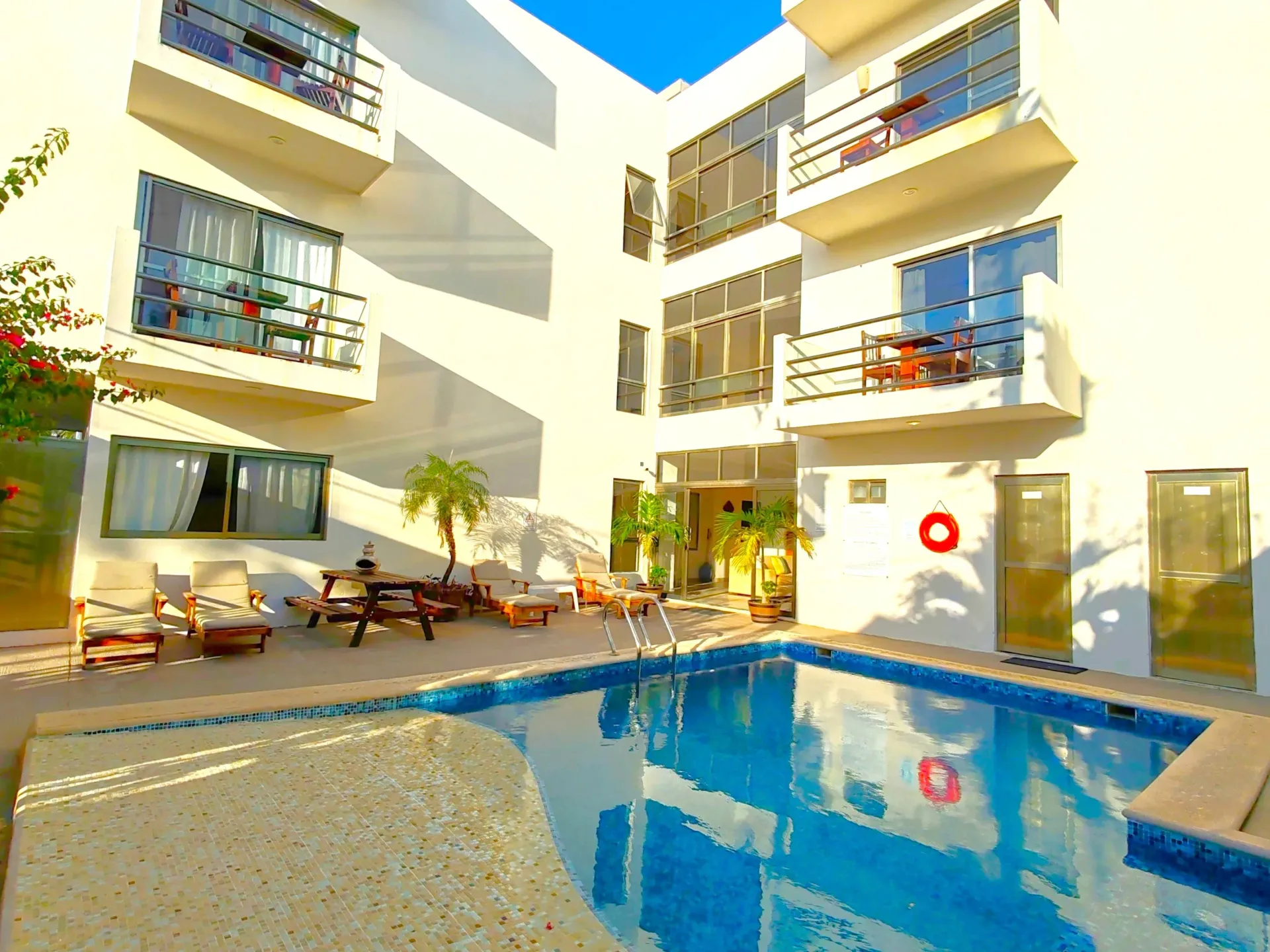 Hotel con patio y piscina, edificio blanco, balcones, tumbonas, agua azul, día soleado.