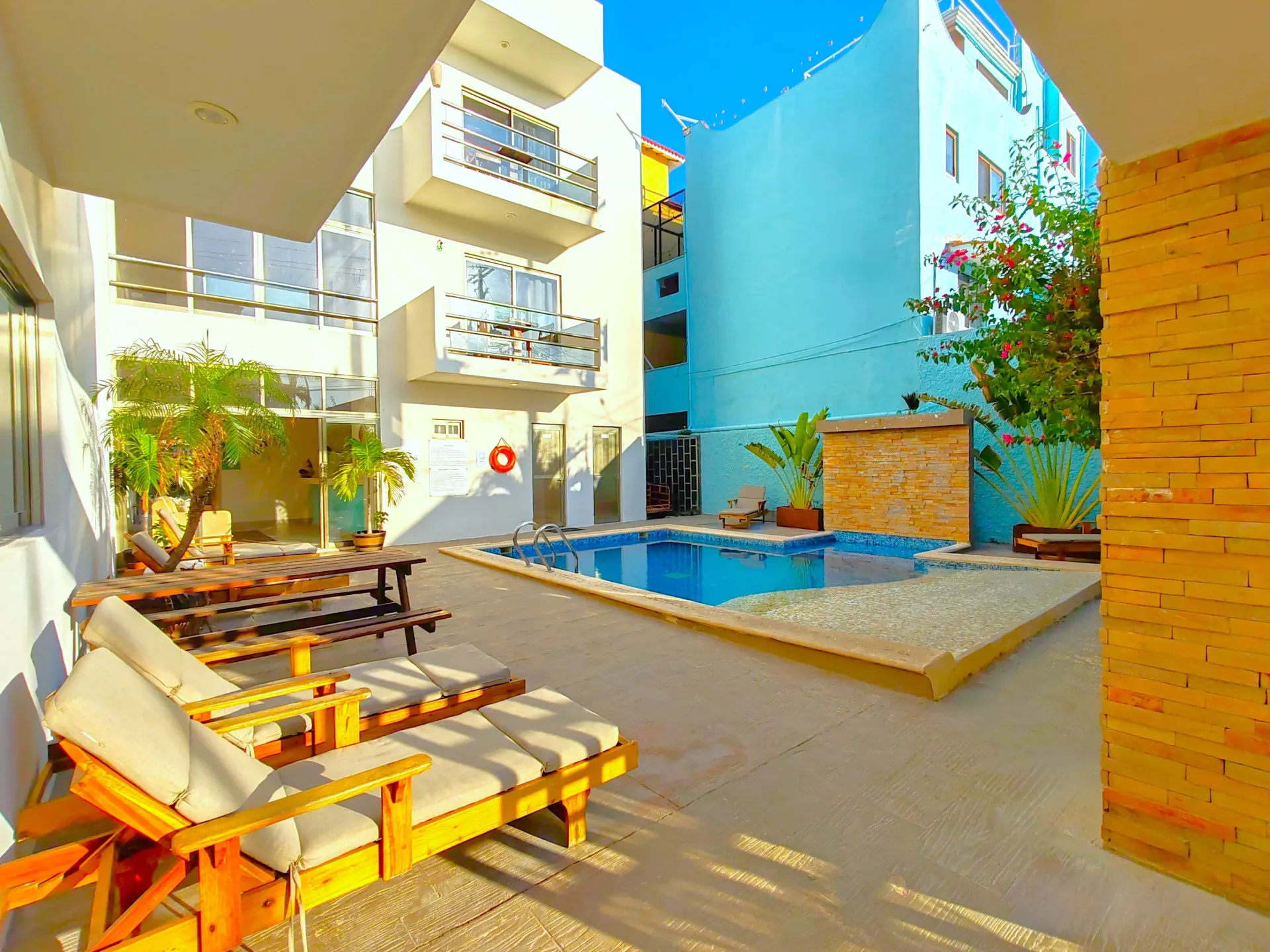 Patio con piscina, tumbonas, mesa de picnic y edificios de varios pisos bajo un cielo soleado.