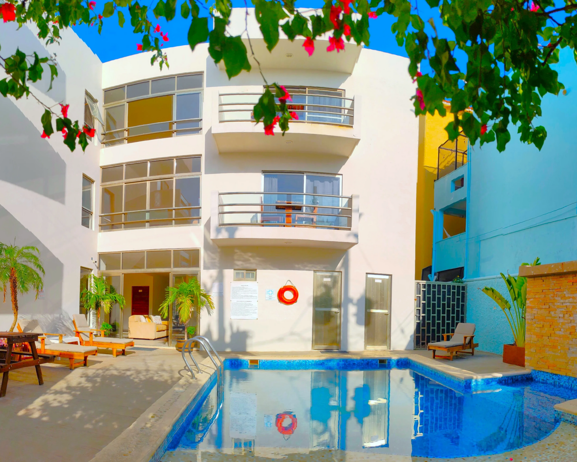 White hotel with pool and lounge chairs, blue sky, and lush greenery.
