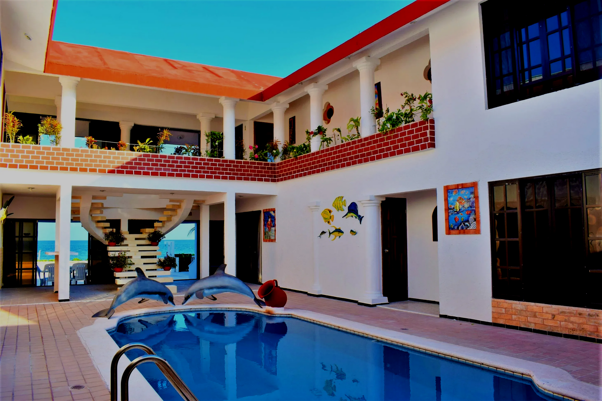 Patio con piscina, edificio blanco, techo rojo, estatuas de delfines, vista al mar.