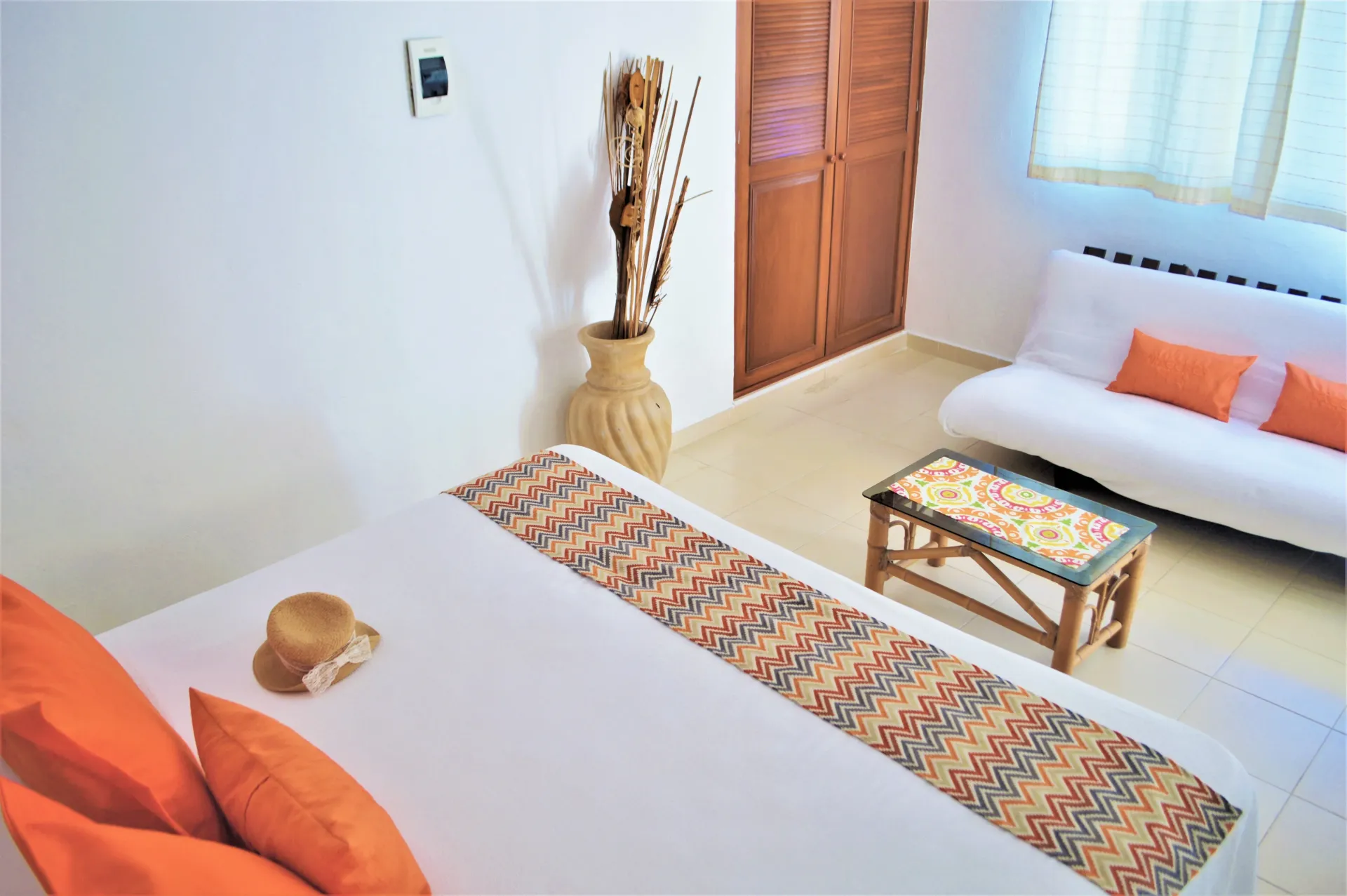 Interior de dormitorio con cama, sofá, mesa y jarrón decorativo. Habitación luminosa, detalles en naranja .