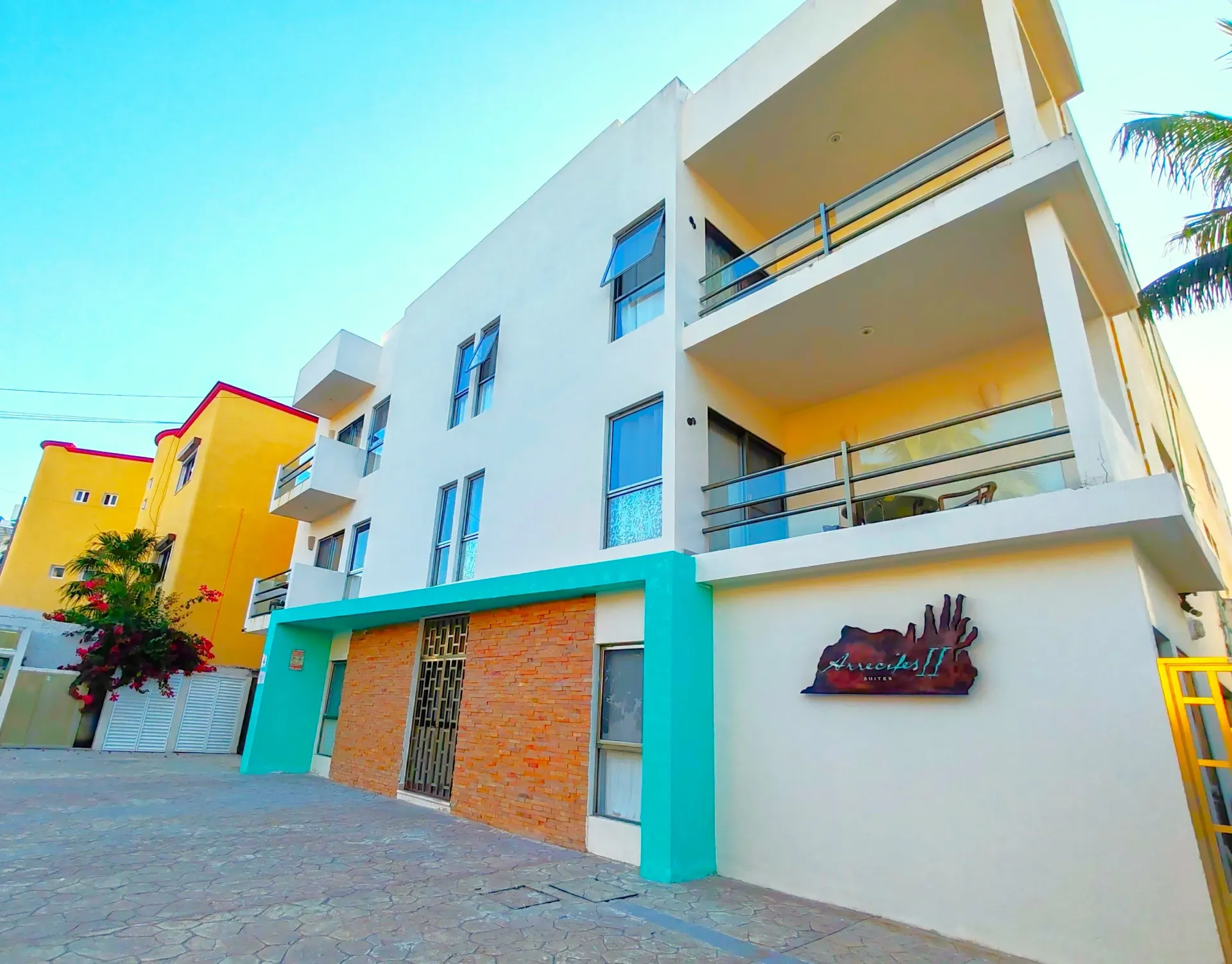 Edificio blanco de varias plantas con balcones, molduras turquesas y detalles de ladrillo. Edificio amarillo al lado.