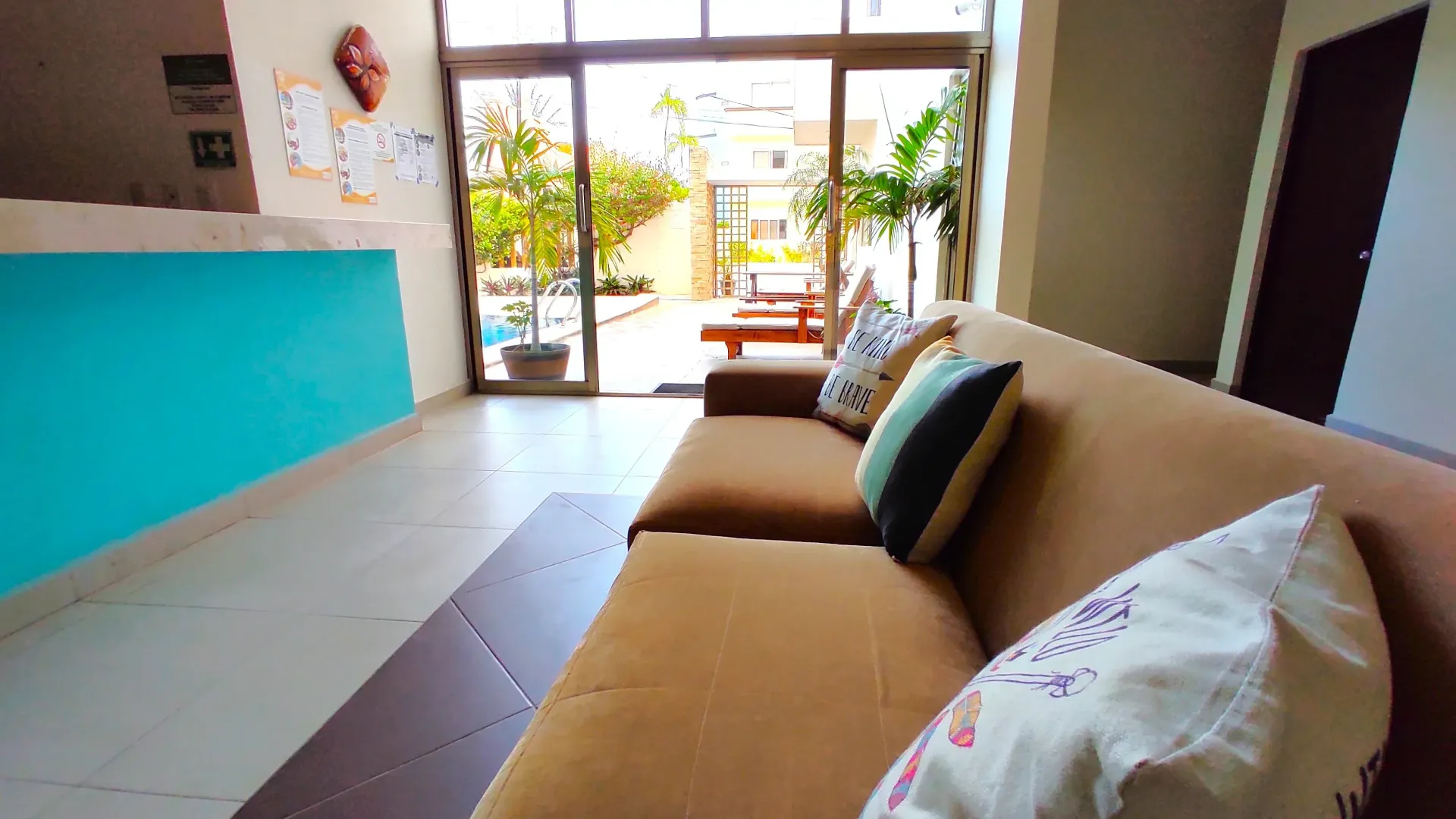 Sofá marrón con almohadones de colores frente a una puerta abierta a un patio, cerca de un mostrador de recepción turquesa.