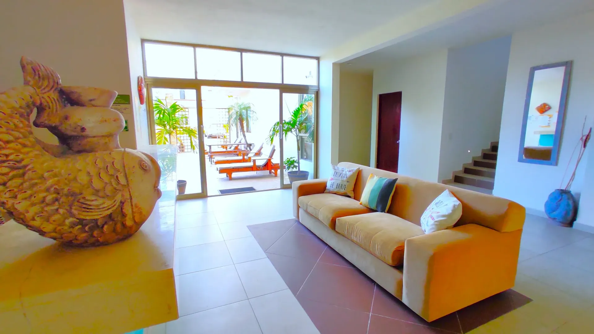 Salón con sofá naranja, escultura y acceso al patio. Tonos neutros y brillantes, con la luz del sol entrando  ventanas.