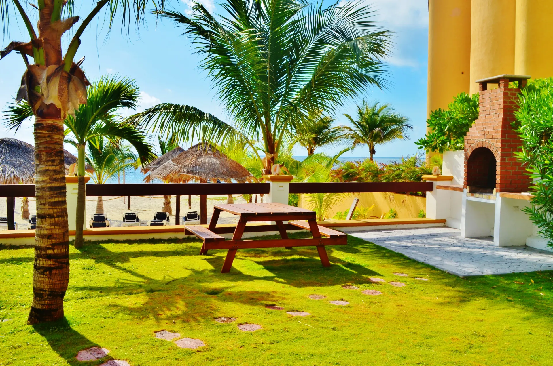 Patio trasero cubierto de césped con mesa de picnic, parrilla de ladrillo y vista al mar detrás de una cerca; día soleado.