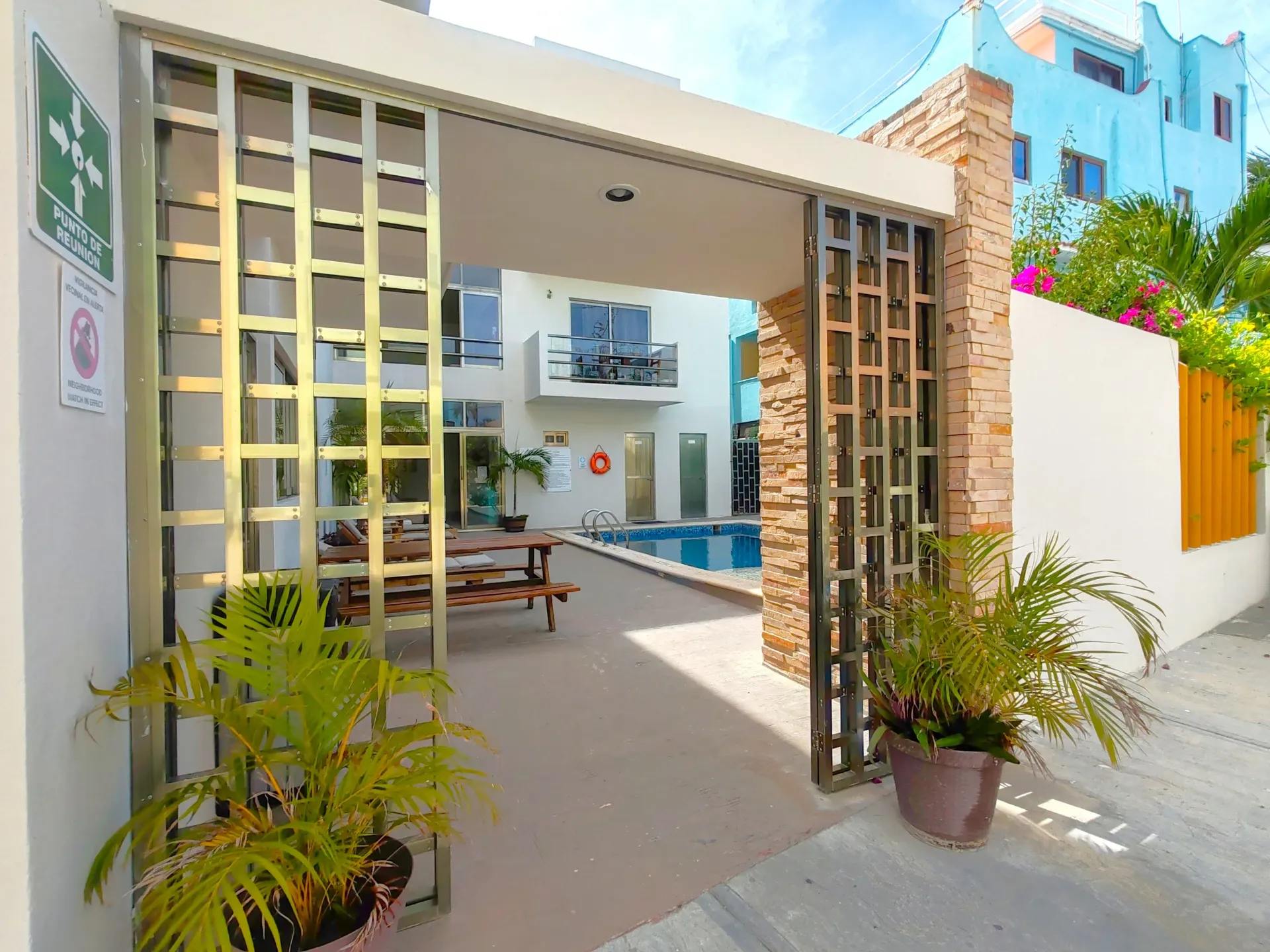 Entrada a un patio con piscina, portón de metal, plantas en macetas y un edificio de color azul claro.