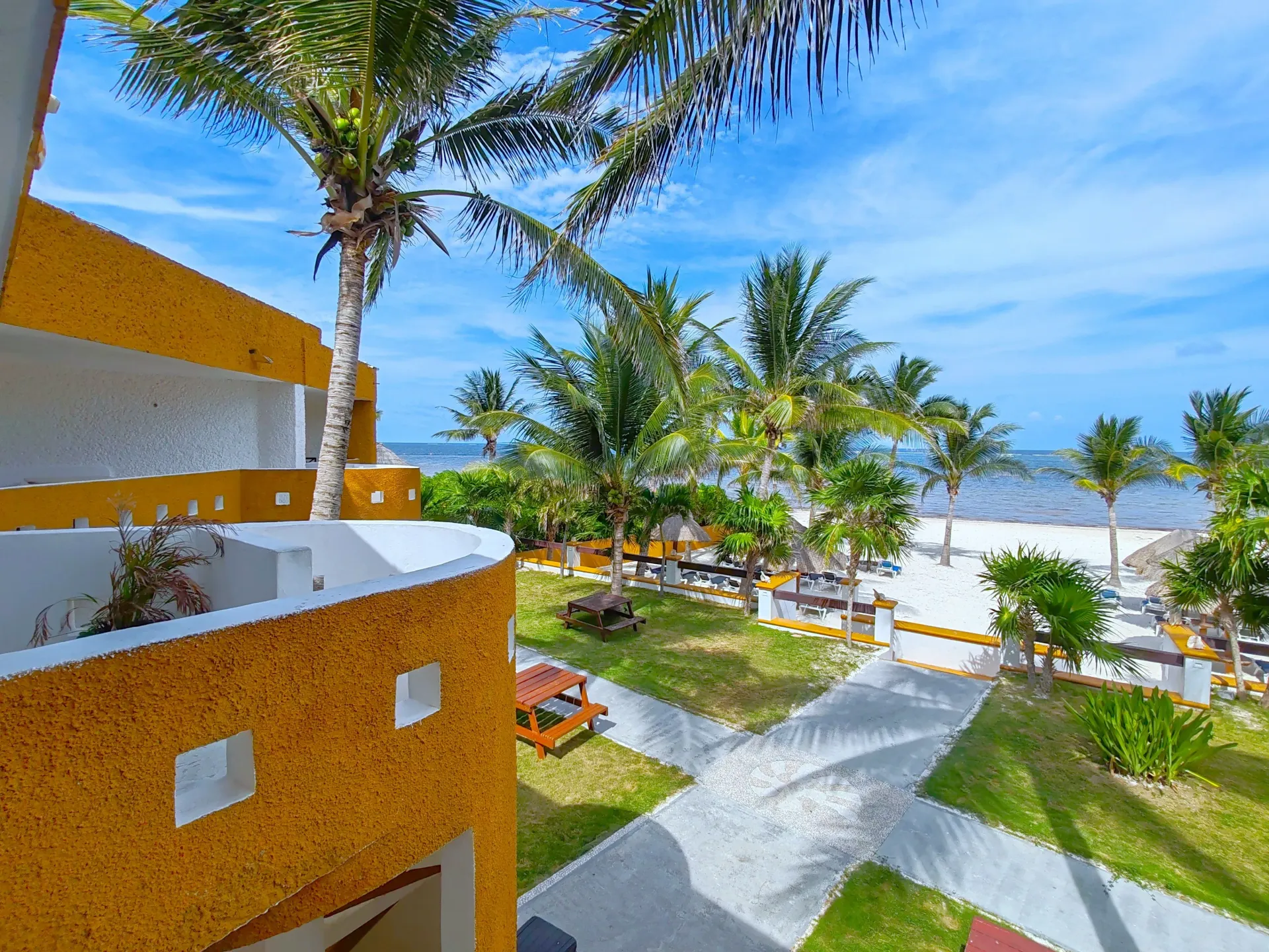 Edificio amarillo con vistas a un patio tropical con palmeras y una playa en un día soleado.