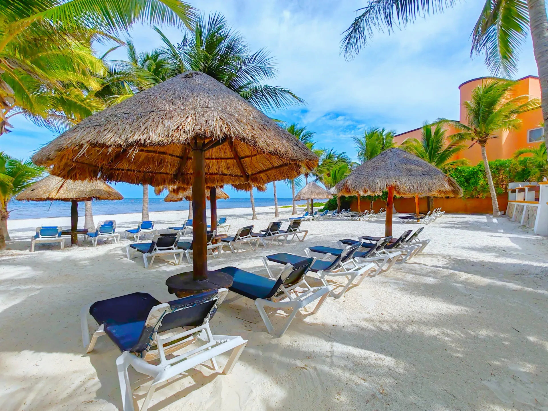 Escena de playa con sombrillas de paja, tumbonas y palmeras en una orilla de arena blanca bajo un cielo azul.