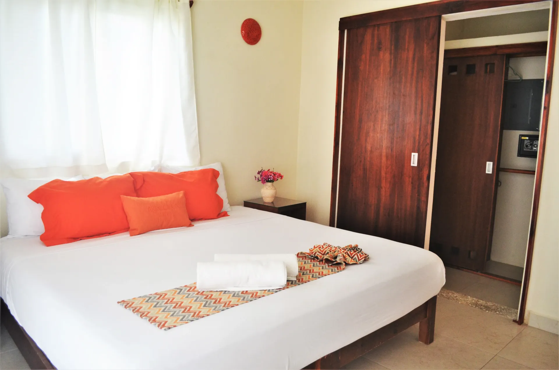 Dormitorio con cama, almohadas naranjas, armario de madera y cortinas blancas.