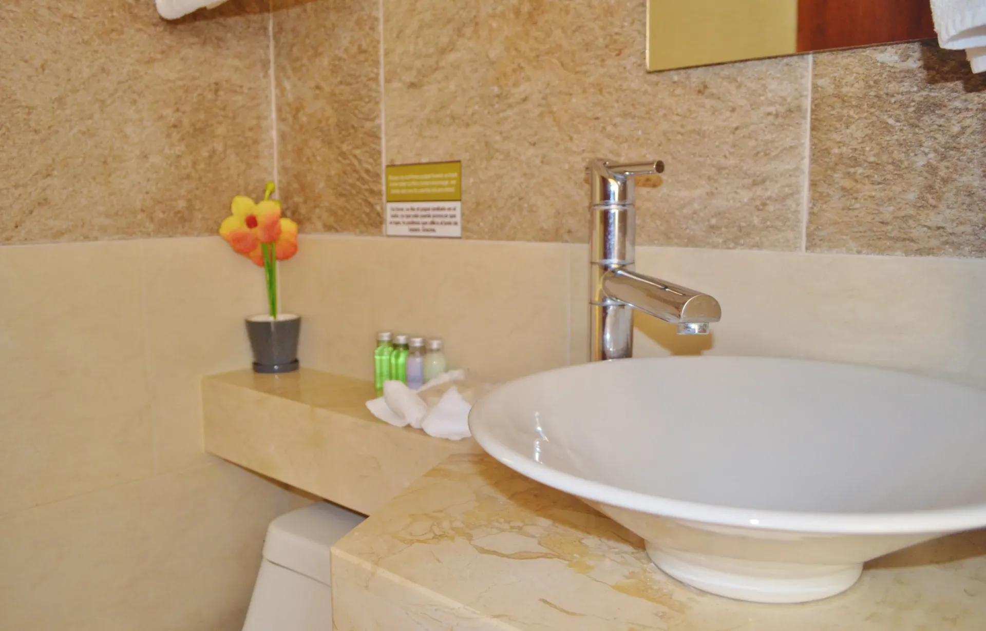 Lavabo de baño con cubeta blanca, grifería cromada, encimera y pared en color beige y una pequeña flor.