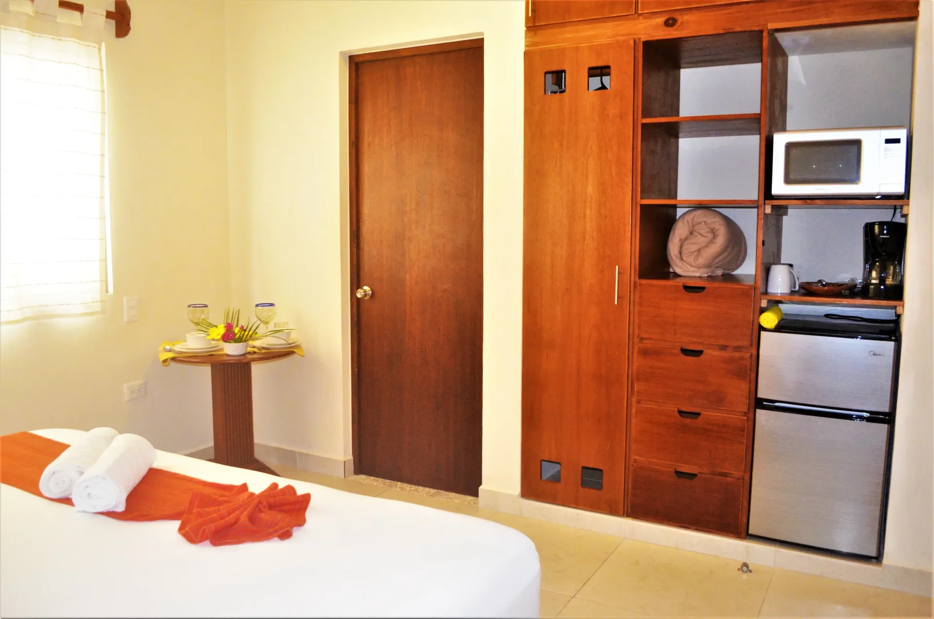 Dormitorio con cama, armario, mininevera, microondas, cafetera y mesita con comida y bebidas.