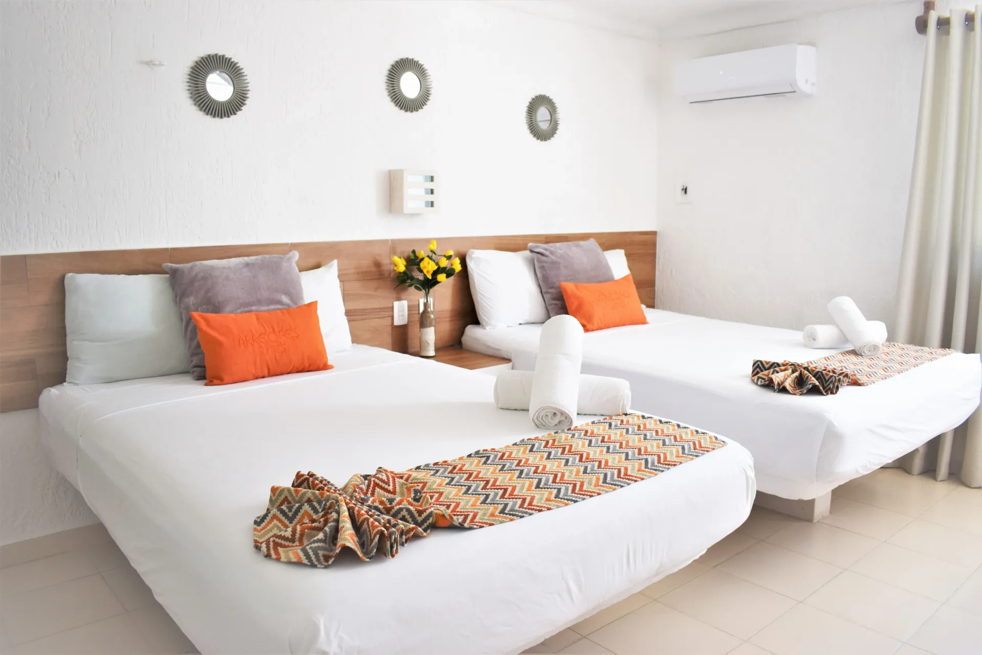 Dormitorio con dos camas, paredes blancas, almohadas naranjas y grises, mantas decorativas.