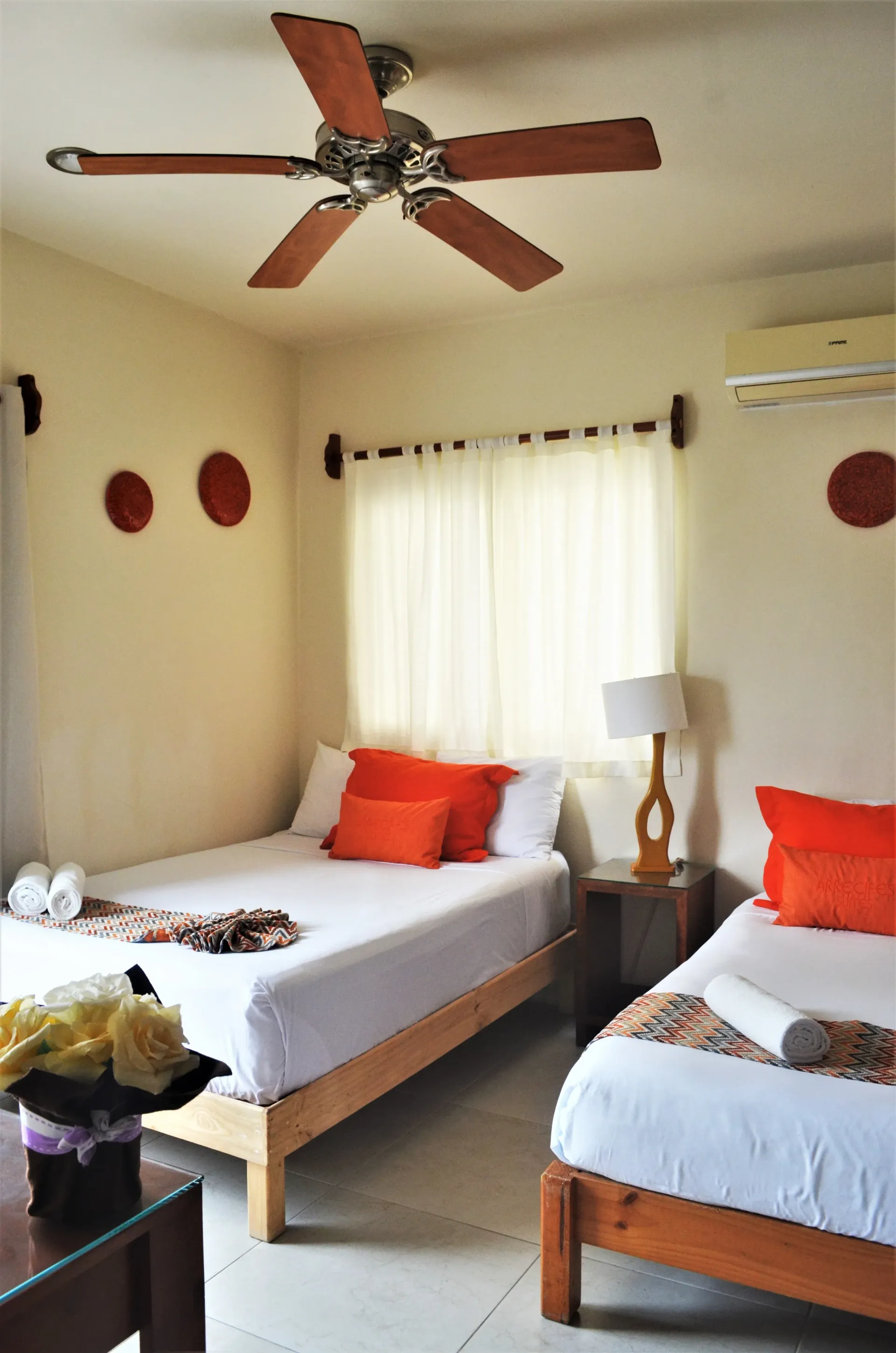 A bedroom with two twin beds, orange pillows, white bedding, and a ceiling fan.