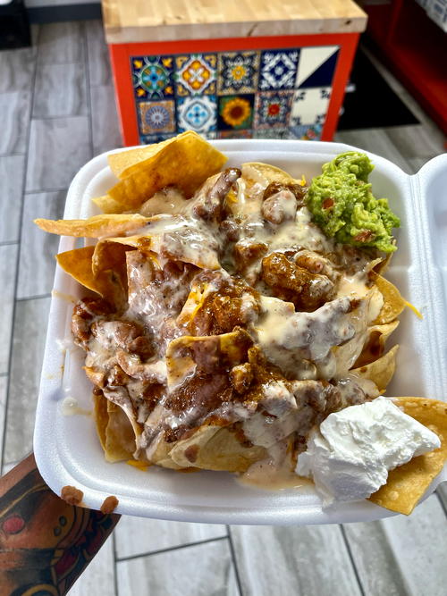 A person is holding a styrofoam container of nachos with guacamole and sour cream.