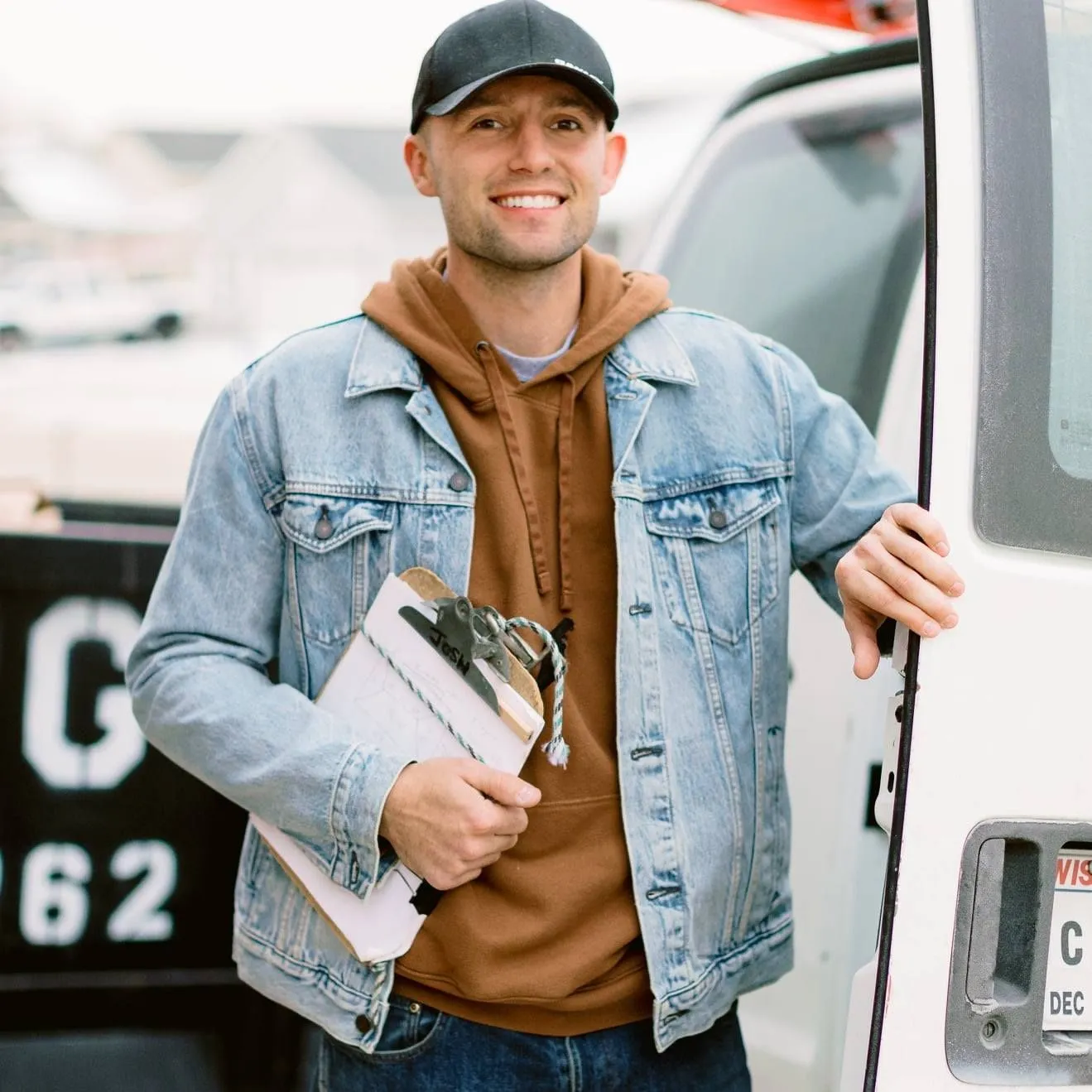 A man in a denim jacket is holding a clipboard in front of a truck with the number 62 on it.