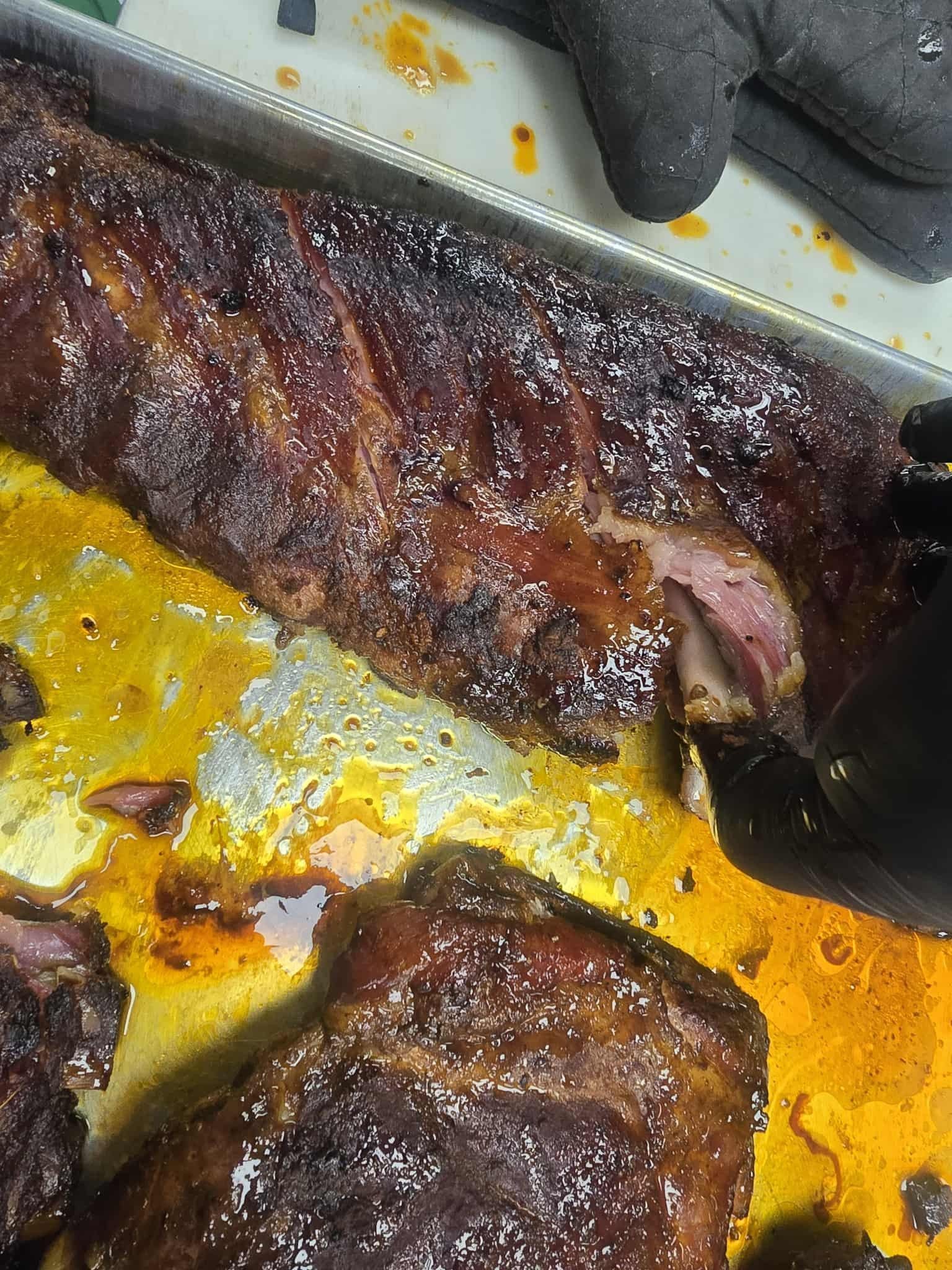 Rack of cooked ribs on a tray; dark brown with glistening glaze and visible bone, being handled with a glove.