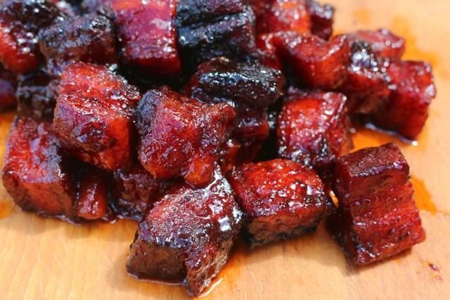 Pile of glistening, deep red-brown pork belly burnt ends, piled on a wooden surface.
