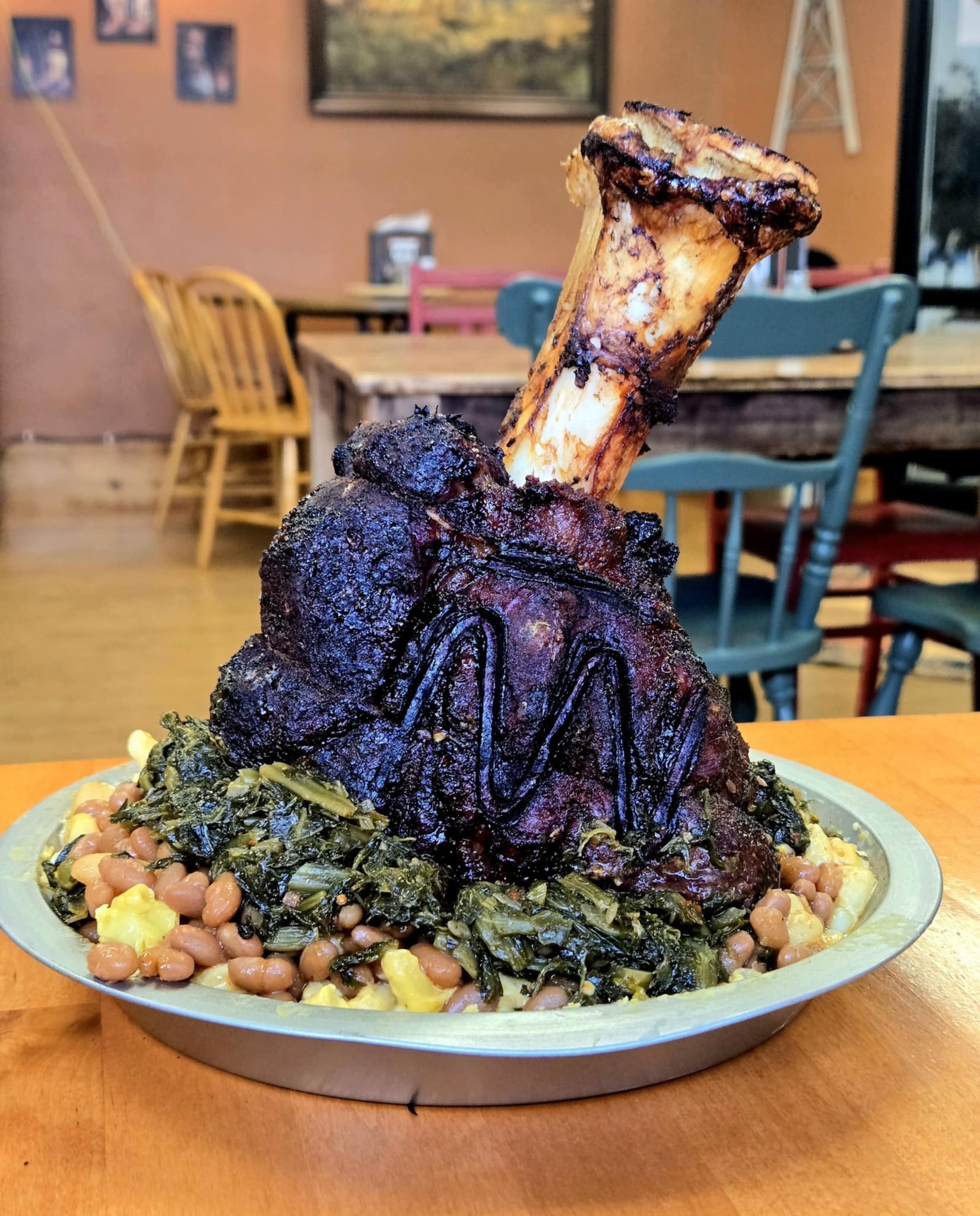 Smoked ham hock atop collard greens and beans on a metal plate in a rustic restaurant.