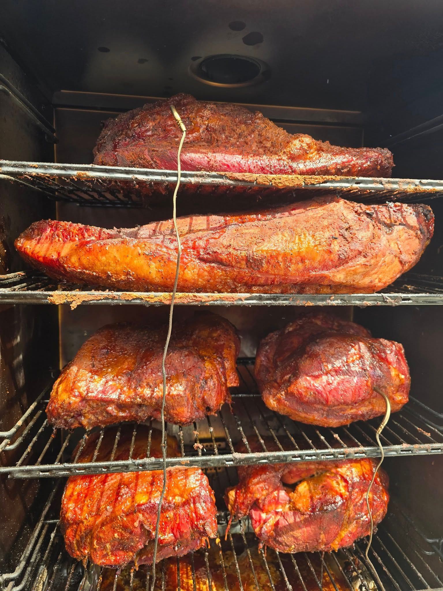 Smoker filled with various cuts of smoked meat, reddish-brown, stacked on racks inside.