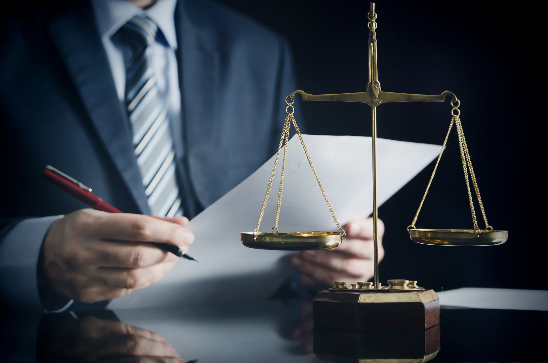 A person in a suit signing documents next to a balanced justice scale on a desk.