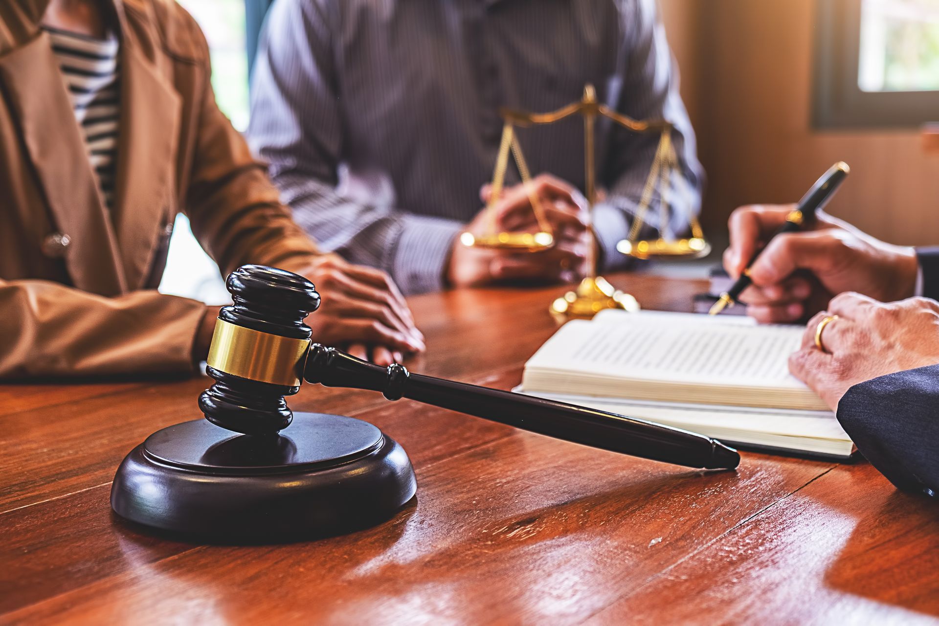 Gavel on table with people discussing legal documents, scales of justice in background.