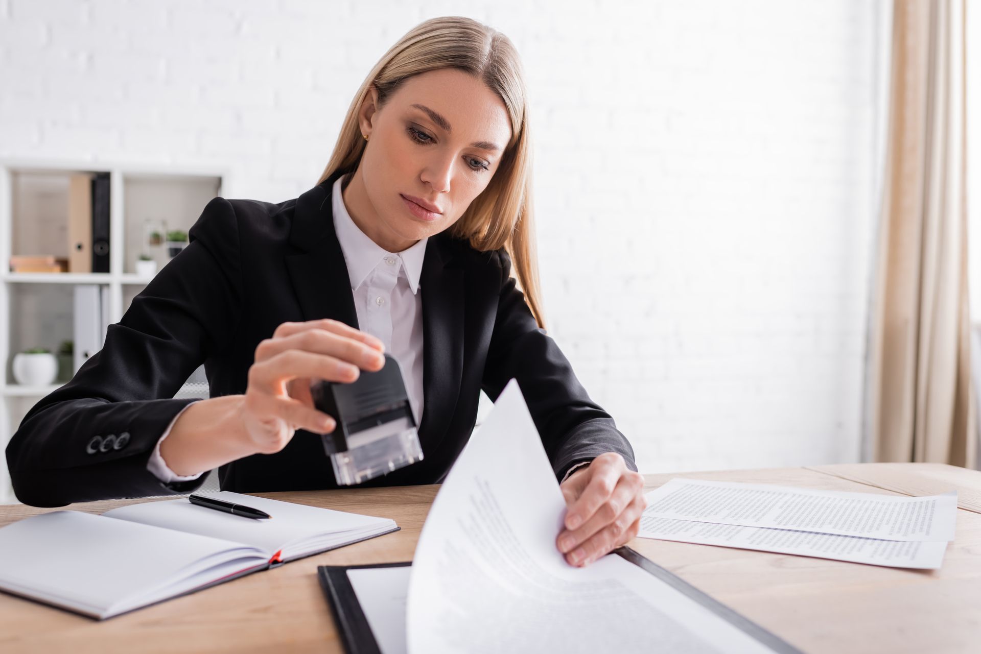 Woman in a blazer stamps a document at a desk with an open notebook, papers, and window in background.