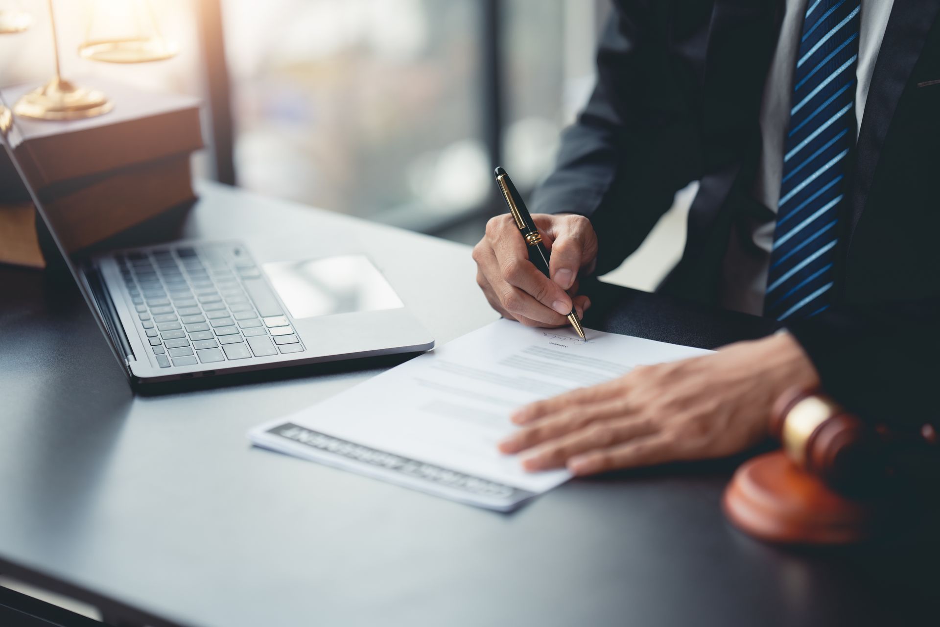 A person in a suit signing legal documents at a desk, laptop and gavel present.