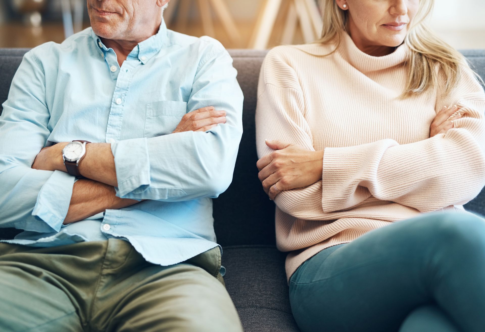 Couple sitting apart on a couch, arms crossed, looking unhappy.