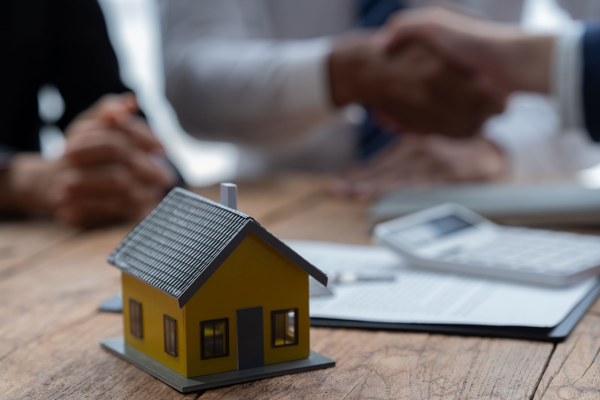 Miniature yellow house on table, with contract, calculator, and two people shaking hands in the background.