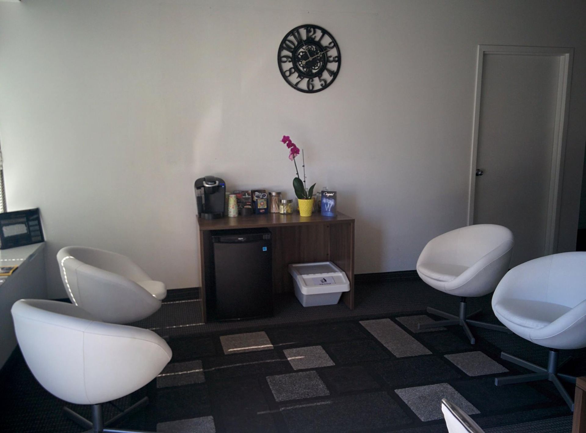 Waiting room with white egg-shaped chairs, coffee station, clock, and a closed white door.