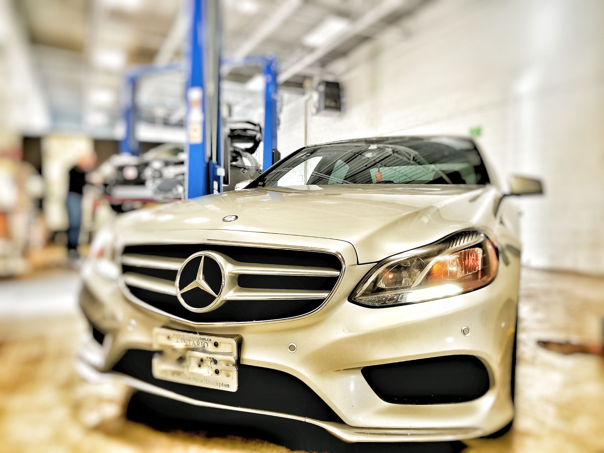 Silver Mercedes-Benz in a repair shop with a mechanic in the background, focused on the car's front. | Mecano Autoworks