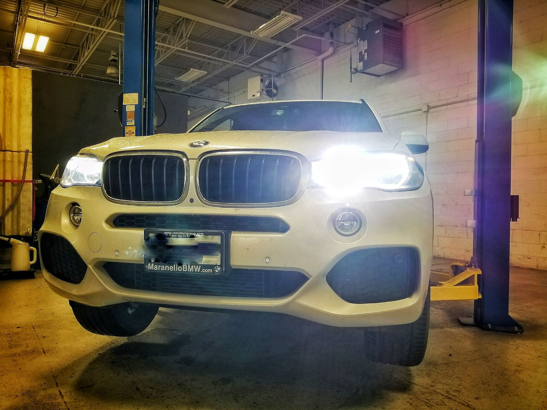White BMW SUV on a hydraulic lift inside a garage, headlights on. | Mecano Autoworks