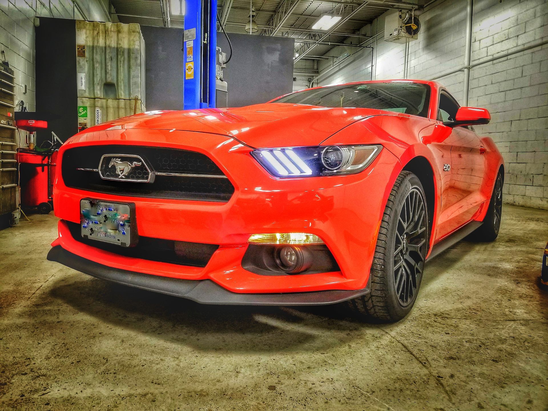 Red Ford Mustang sports car in an auto shop, black wheels, black front spoiler, and blue car lift.
