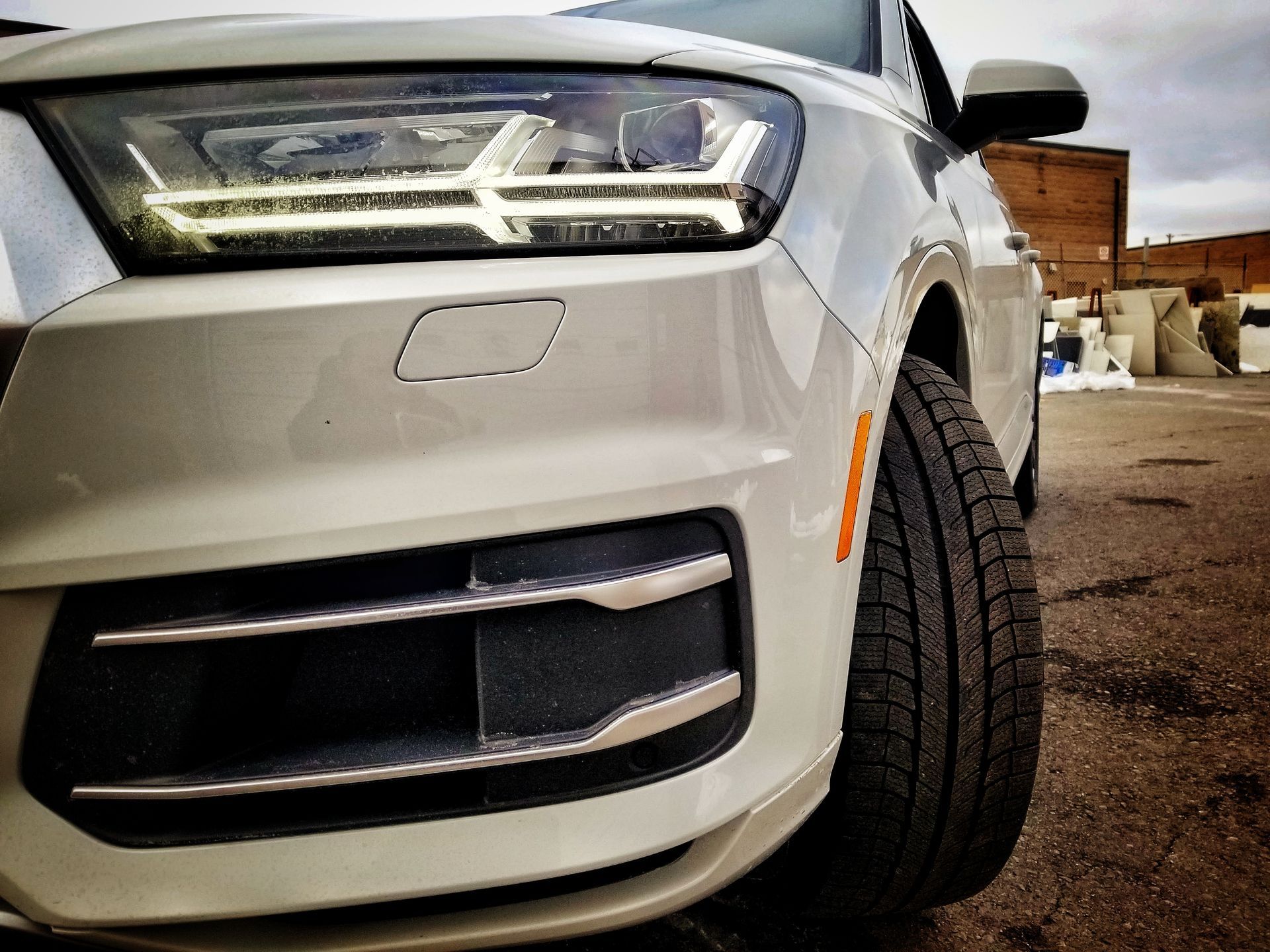 White Audi SUV, close-up view of front, showing headlight, tire, and lower grill. Daytime, outdoors, industrial setting.