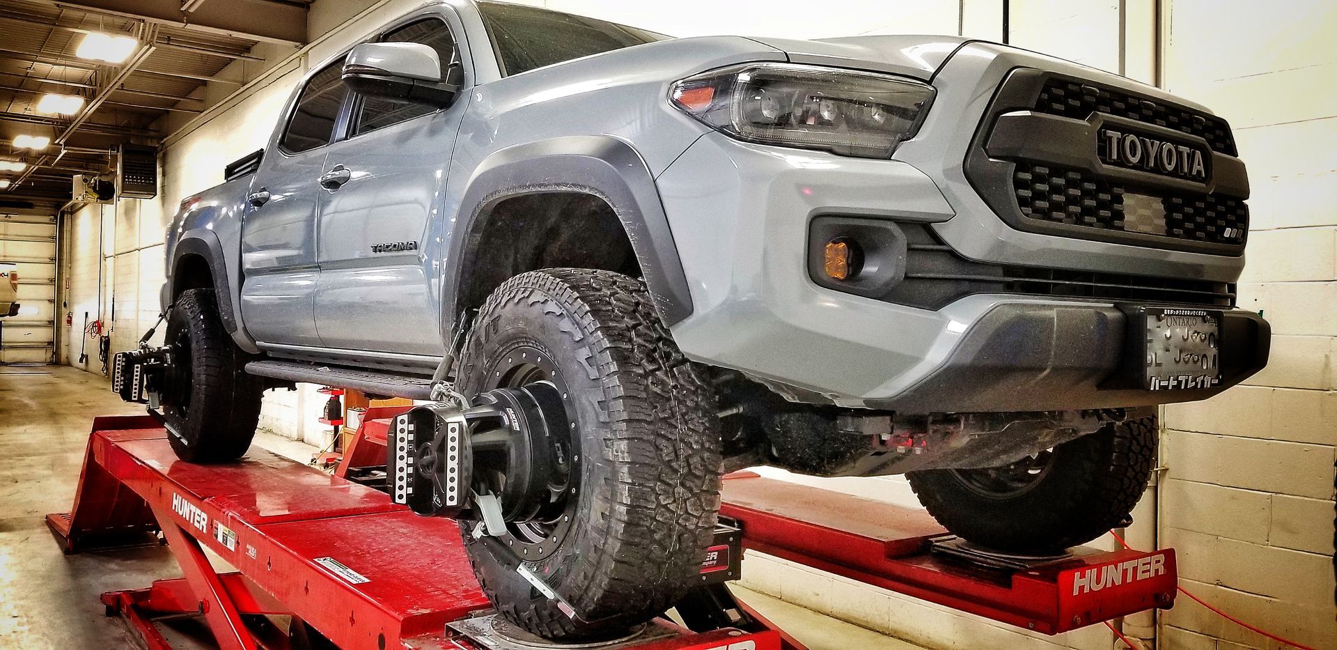 A gray Toyota Tacoma truck is on a red hydraulic lift in a garage, getting a wheel alignment. | Mecano Autoworks