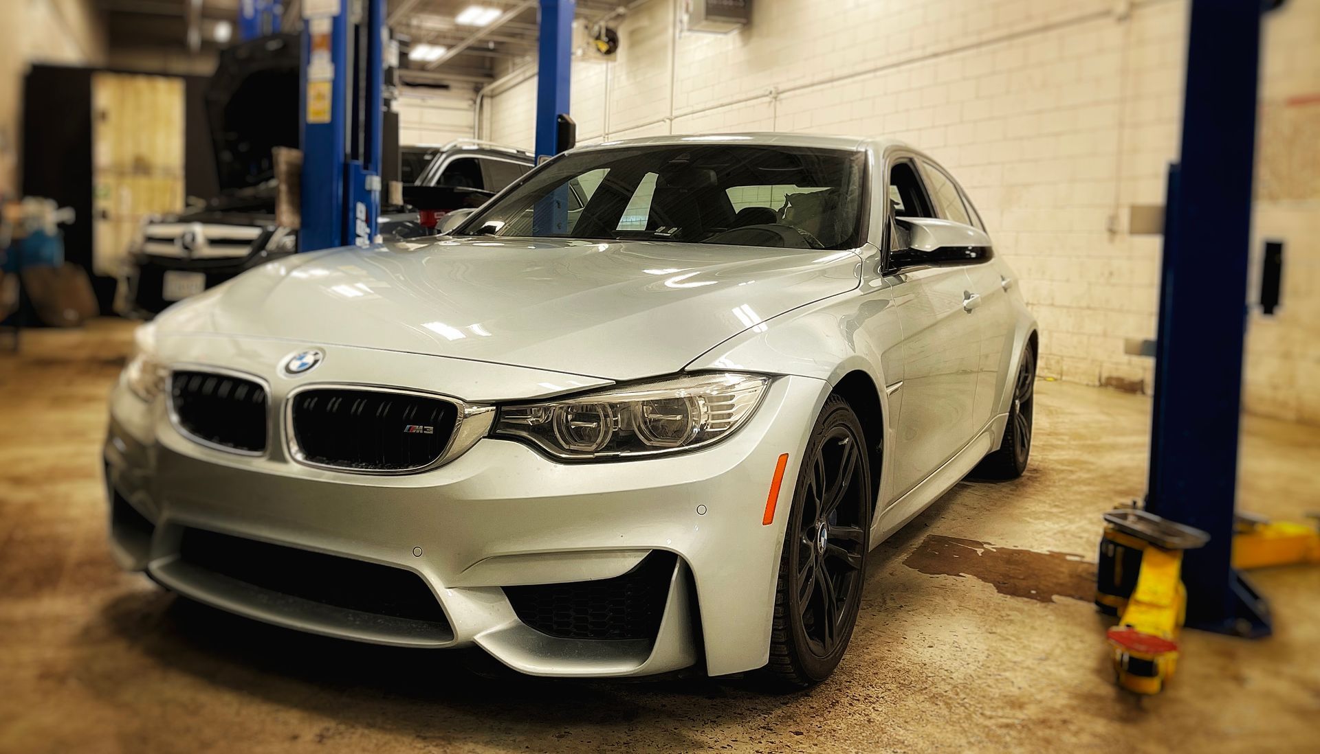 Silver BMW M3 sedan in a mechanic's shop, with blue lift columns, likely for service. | Mecano Autoworks