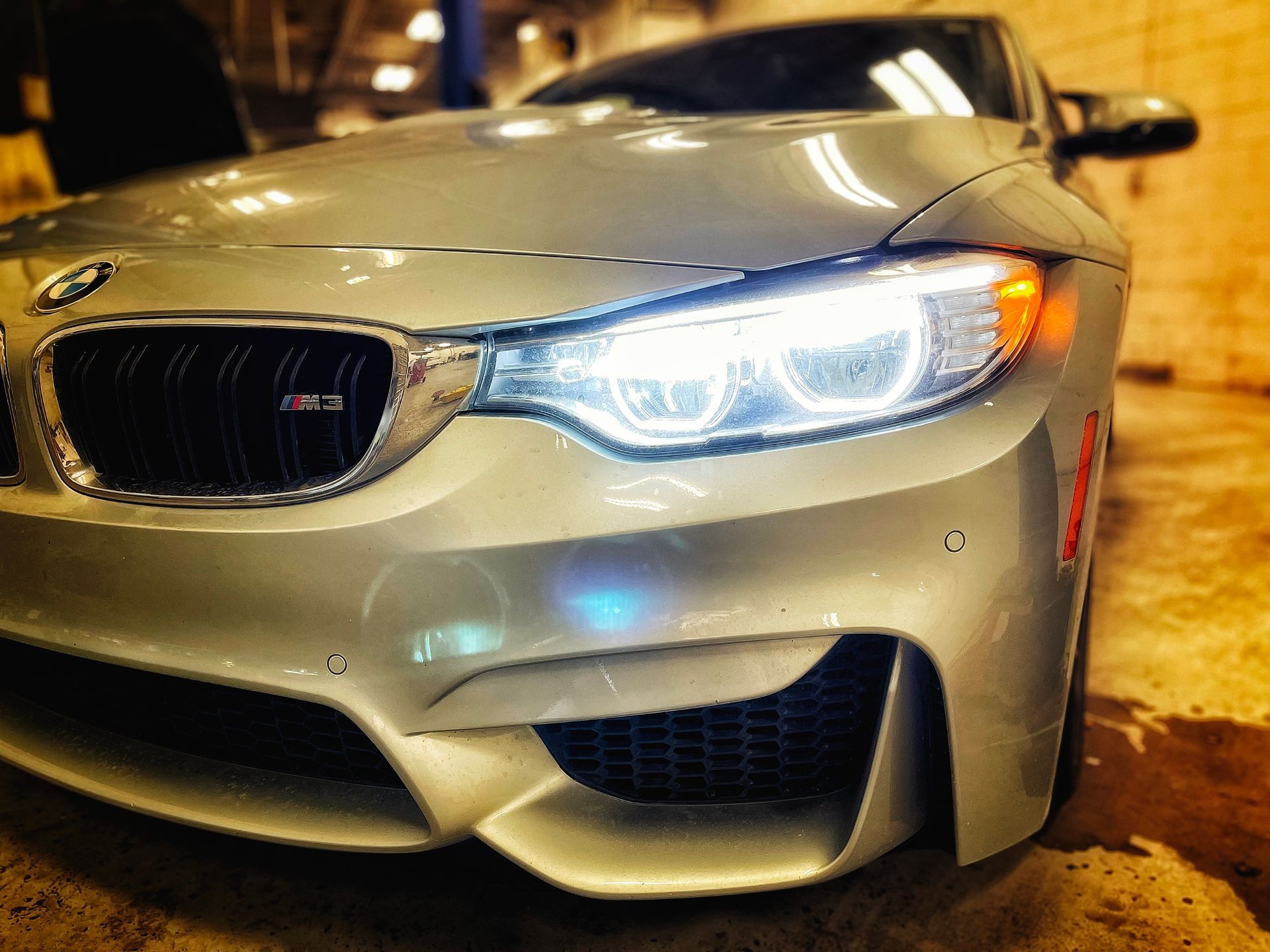 Silver BMW M4 sports car with bright headlights, parked inside a garage. | Mecano Autoworks