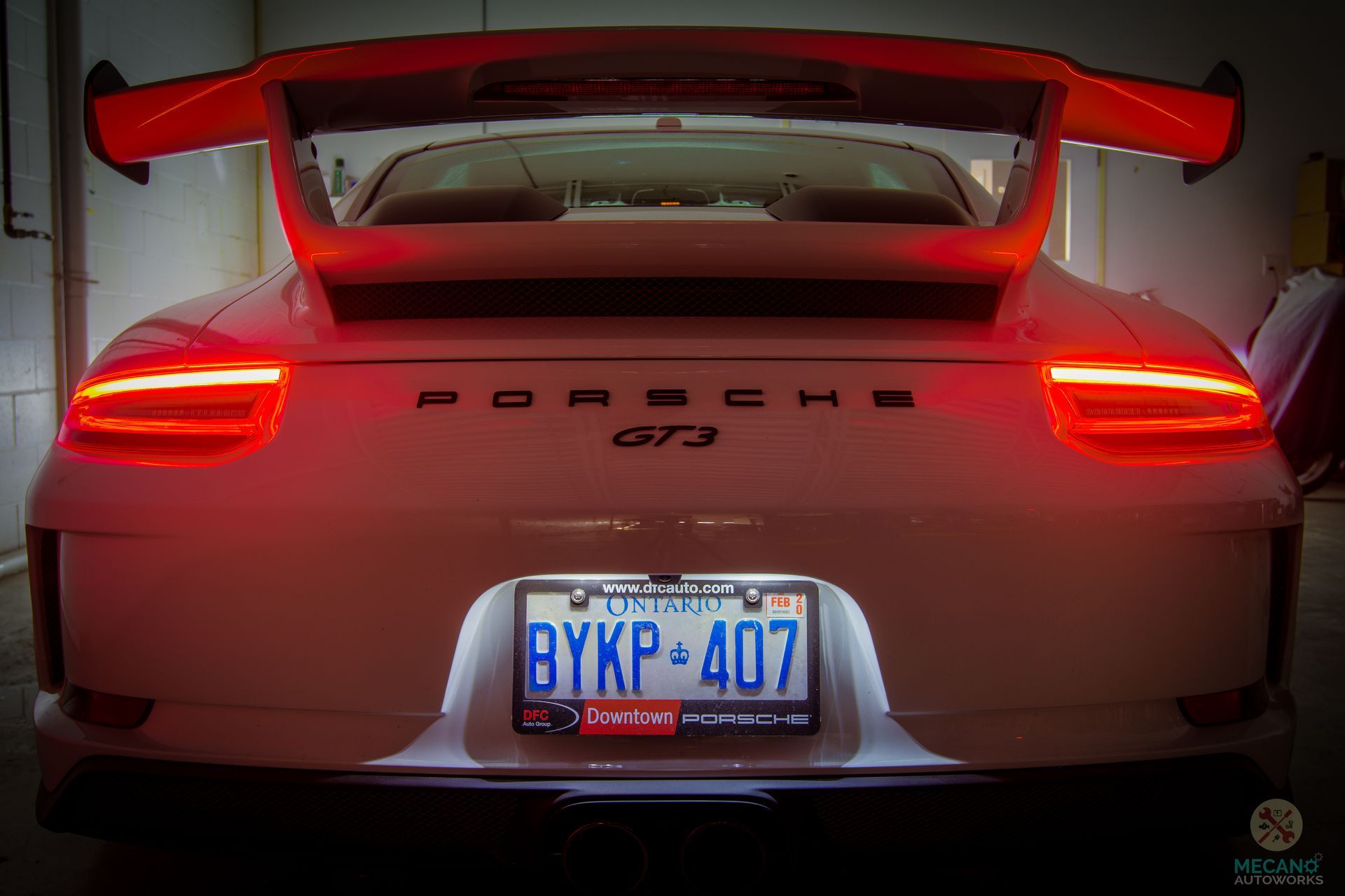 White Porsche GT3 from the rear, lit taillights, large orange wing, license plate, dark background. | Mecano Autoworks